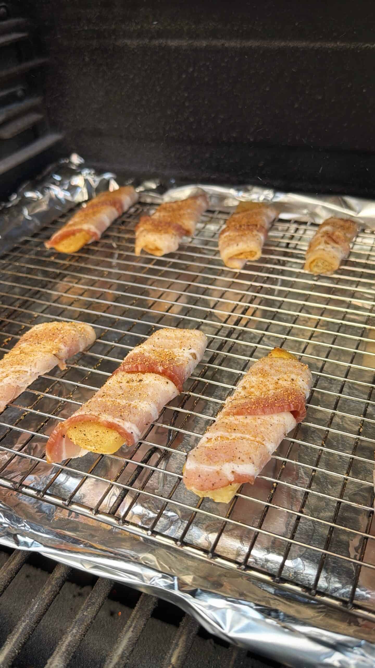 Several bacon-wrapped food items, seasoned and uncooked, are arranged on a wire rack set over a foil-covered baking sheet inside an oven or grill.