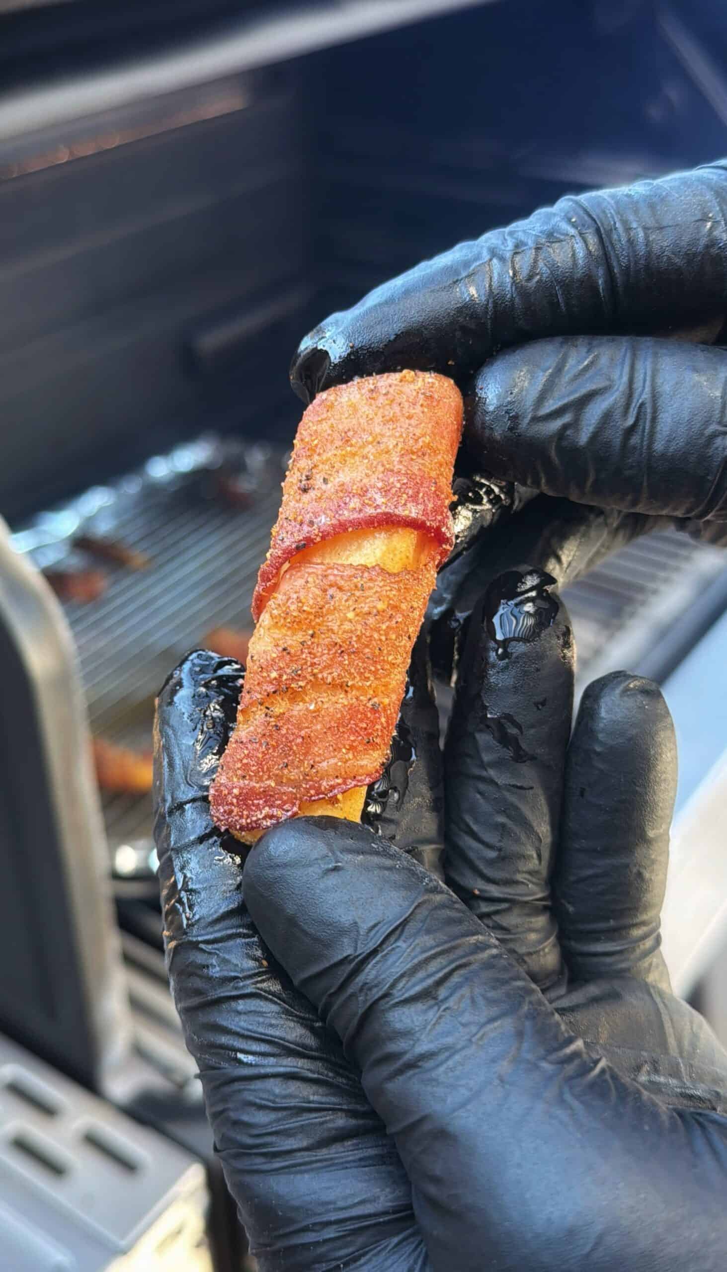 A close-up of gloved hands holding a seasoned, bacon-wrapped food item in front of a grill. The bacon appears crispy and well-cooked, with a grill grate and more food in the background.
