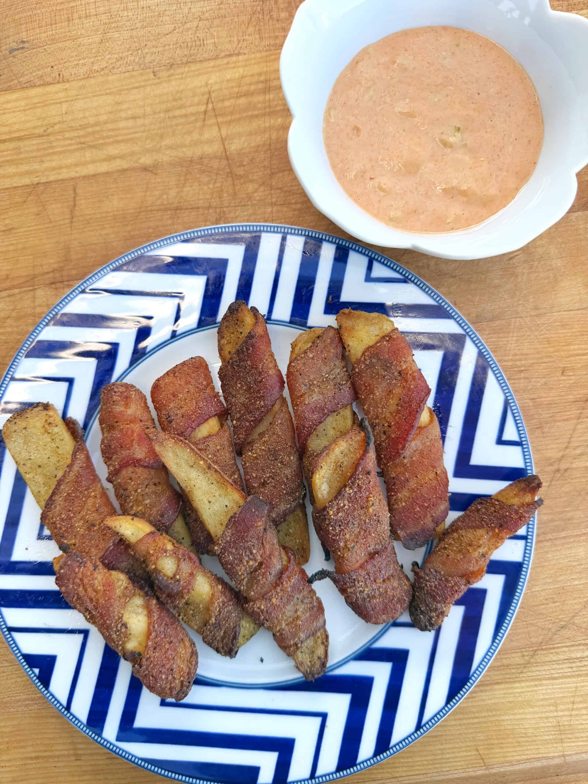 A plate of potato wedges wrapped in bacon, arranged on a blue and white patterned plate, sits on a wooden surface next to a bowl of creamy dipping sauce.
