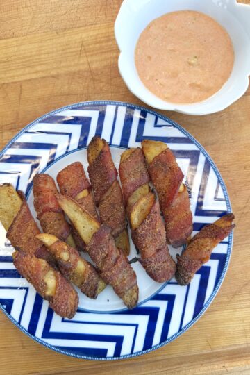 A plate of bacon-wrapped potato wedges arranged on a blue and white patterned plate, next to a bowl of creamy dipping sauce, all set on a wooden surface.