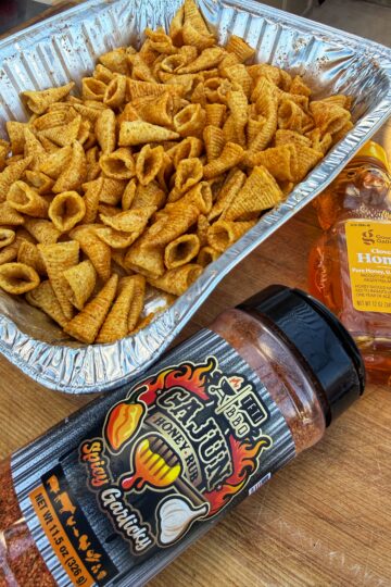 A foil tray filled with Bugles snacks sits on a wooden table next to a bottle of honey and a large container of Cajun honey garlic spicy seasoning.
