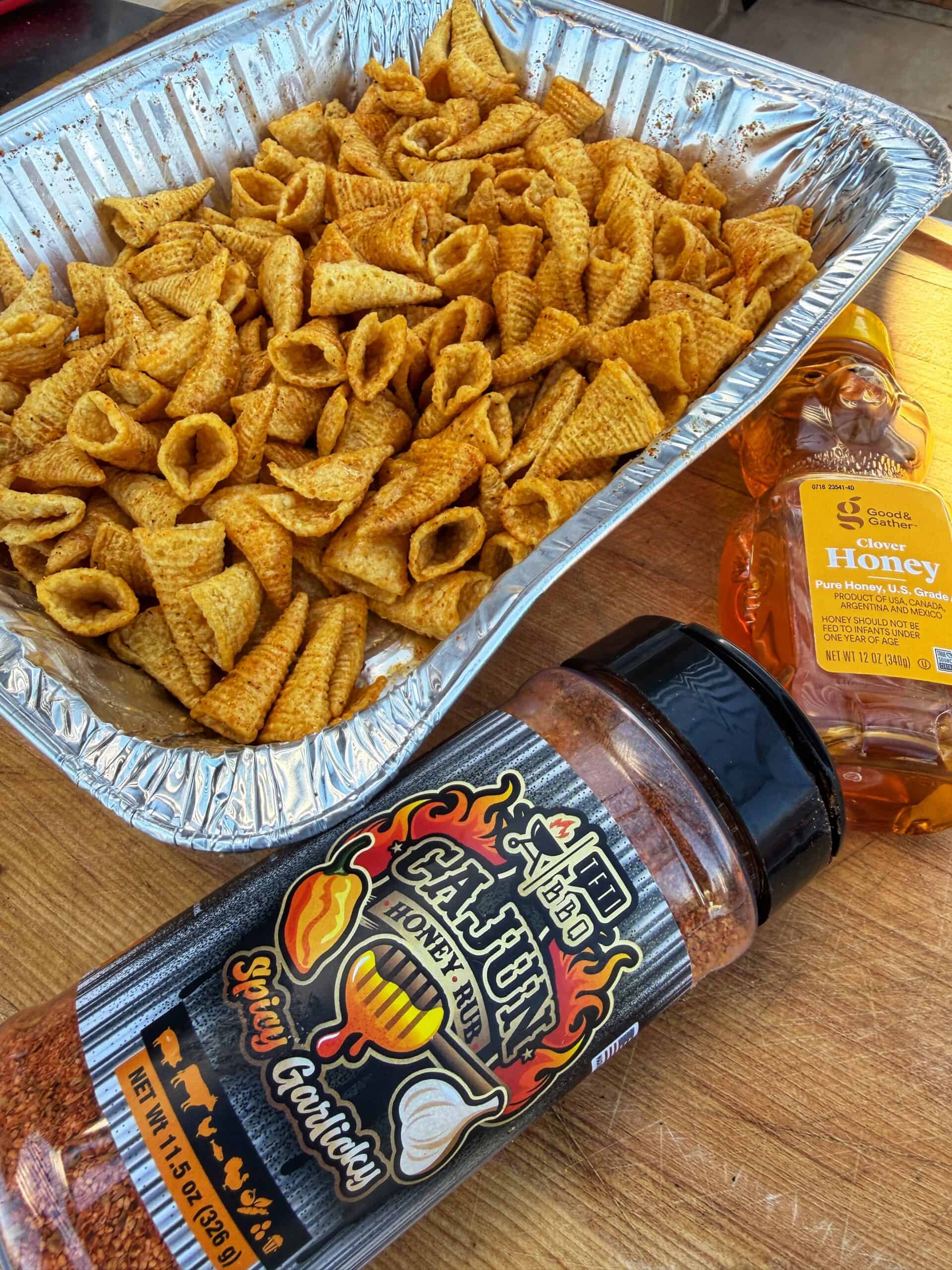 A foil tray filled with Bugles snacks sits on a wooden table next to a bottle of honey and a large container of Cajun honey garlic spicy seasoning.