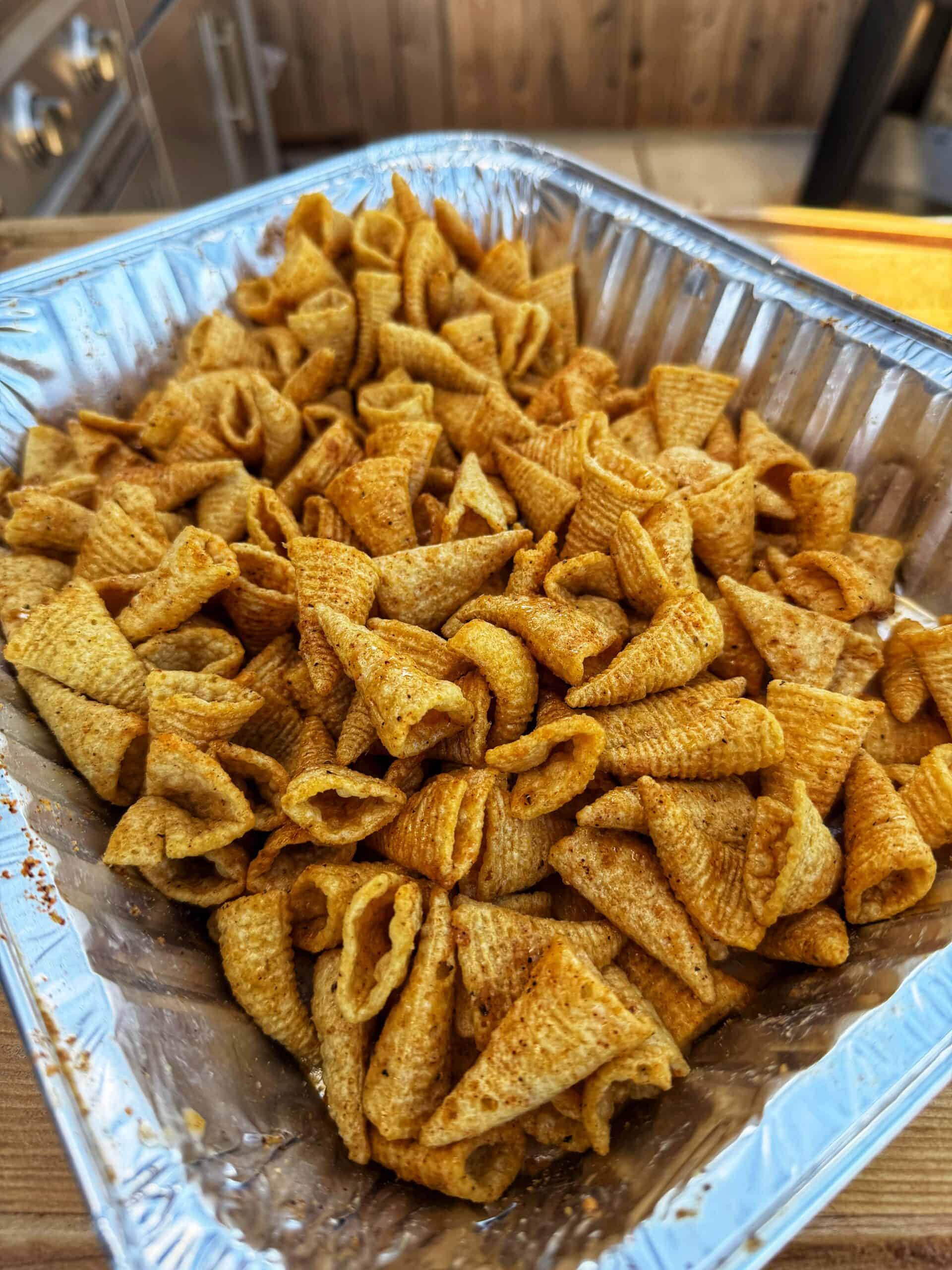 A large aluminum tray filled with crunchy, seasoned corn snacks, resembling Bugles chips, sits on a wooden counter in an outdoor kitchen setting.