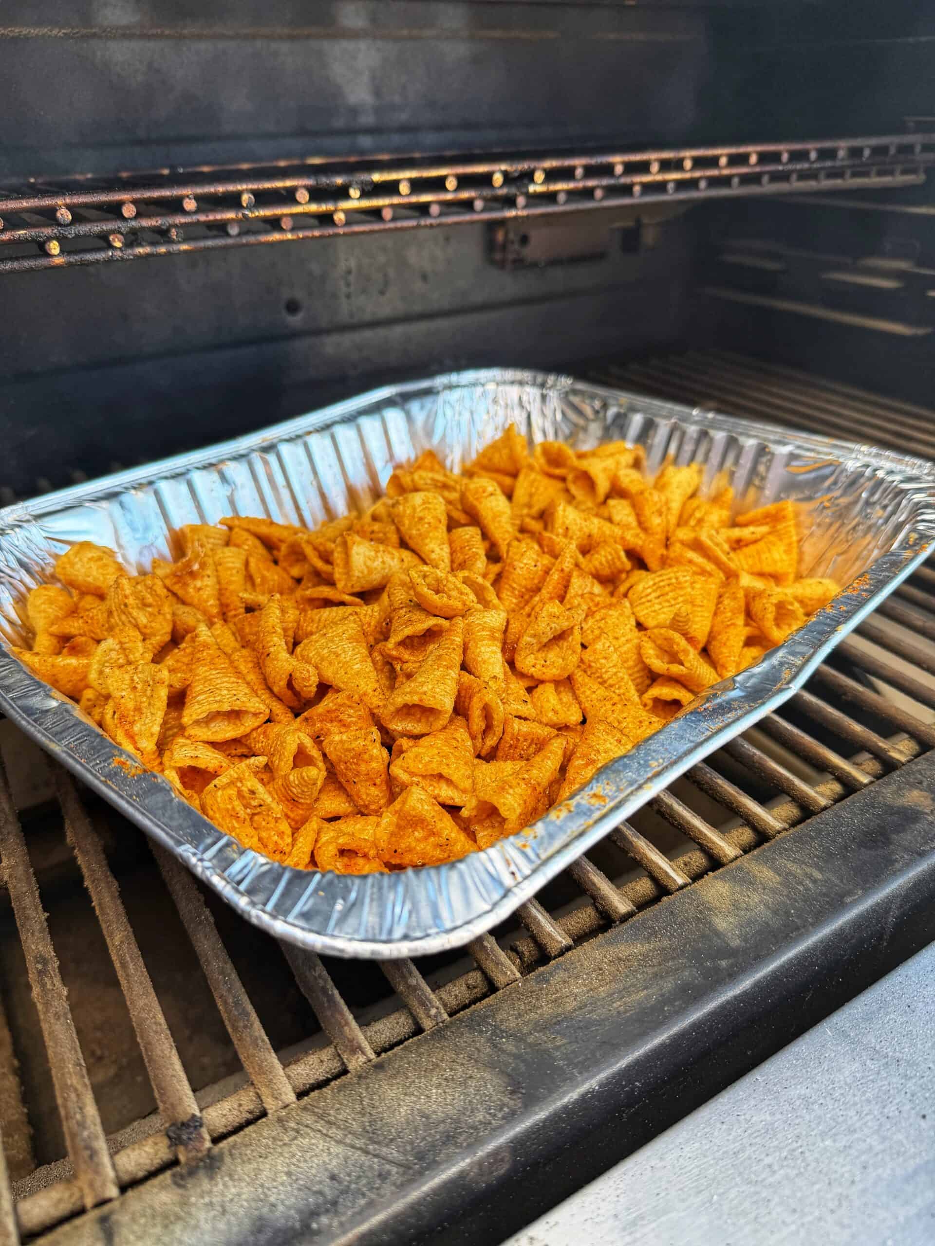 A disposable aluminum tray filled with seasoned corn chips sits on the rack inside a barbecue grill, ready to be smoked or cooked.