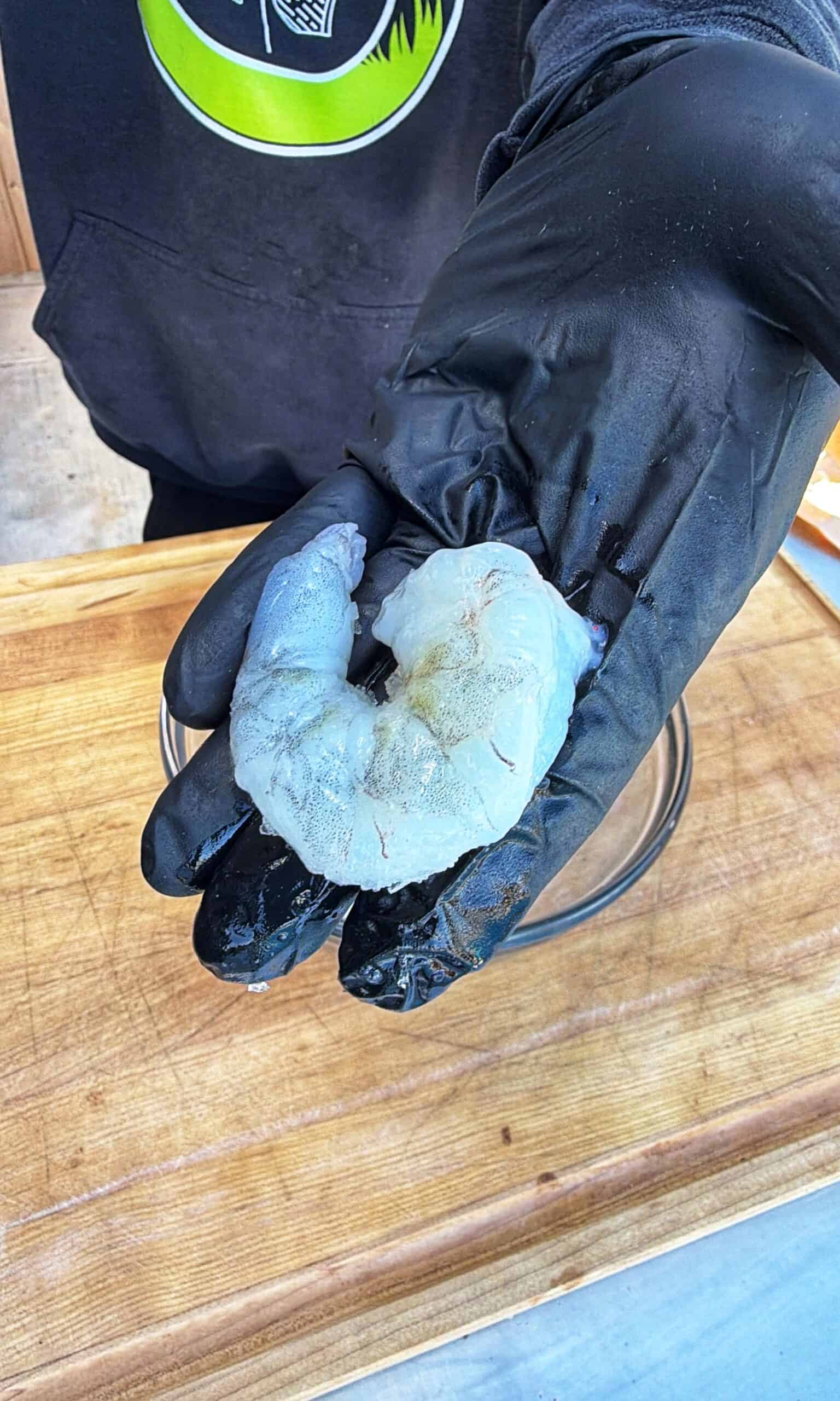 A person wearing a black glove holds a large raw shrimp over a wooden cutting board.