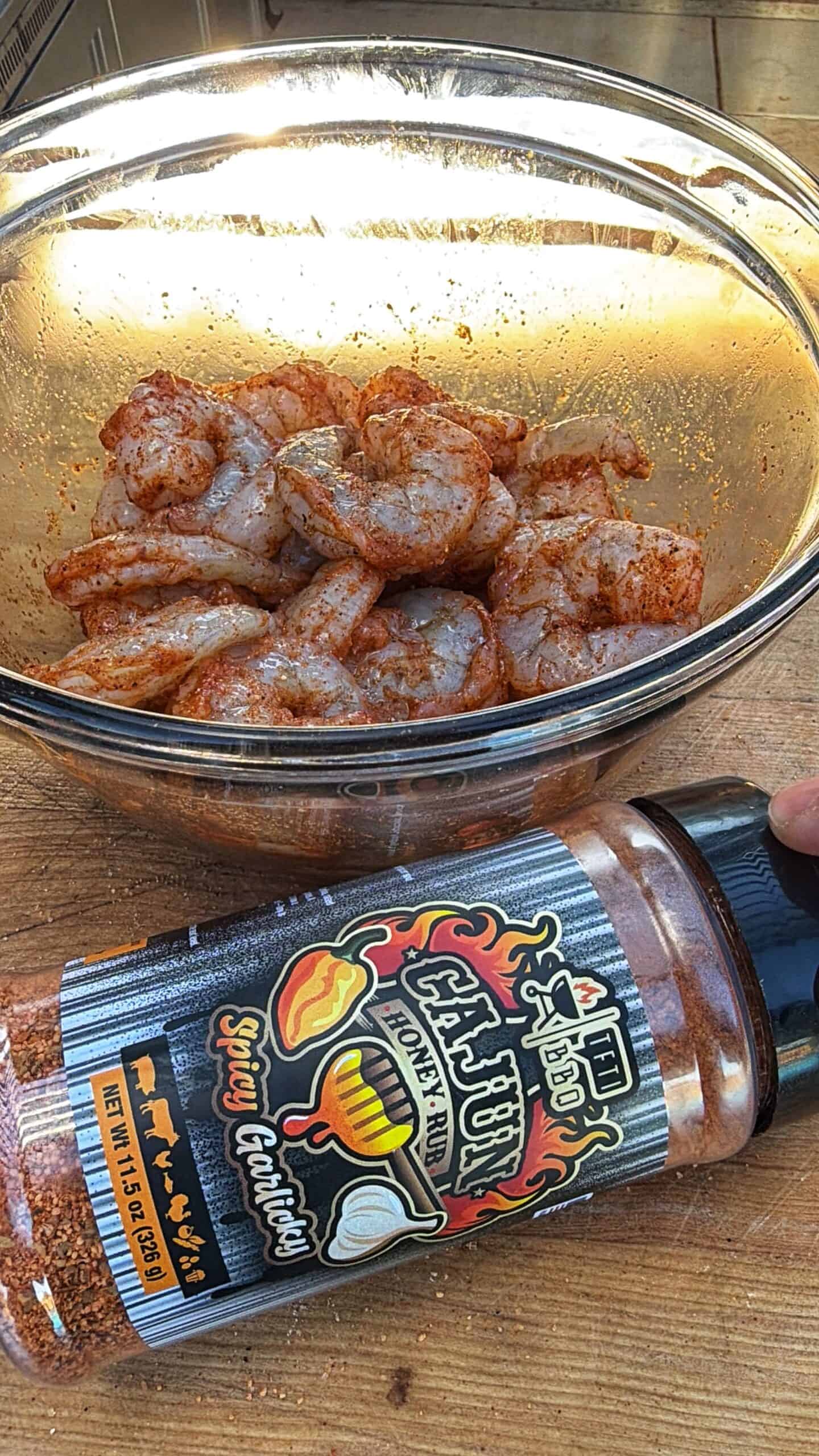 A glass bowl filled with raw shrimp coated in seasoning sits on a wooden surface. In the foreground, a bottle of Cajun spice labeled Spicy Garlic is held by a hand.