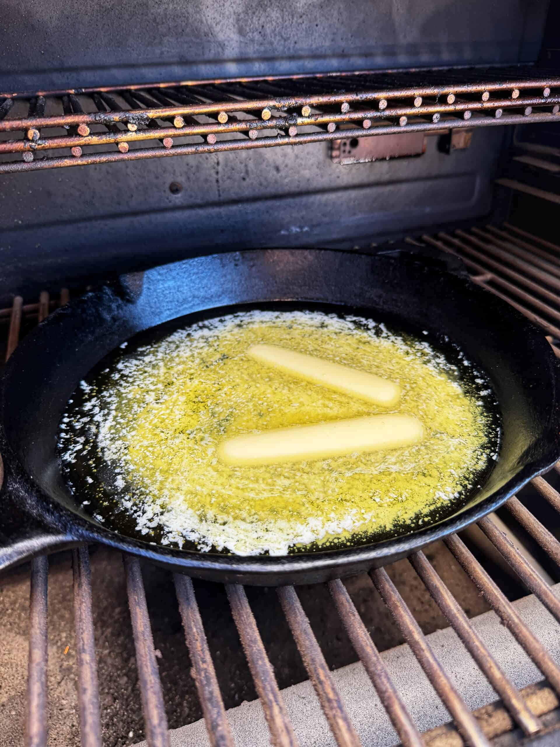 Two sticks of butter melting in a black cast iron skillet on a grill, with the butter forming a greater than symbol as it melts.