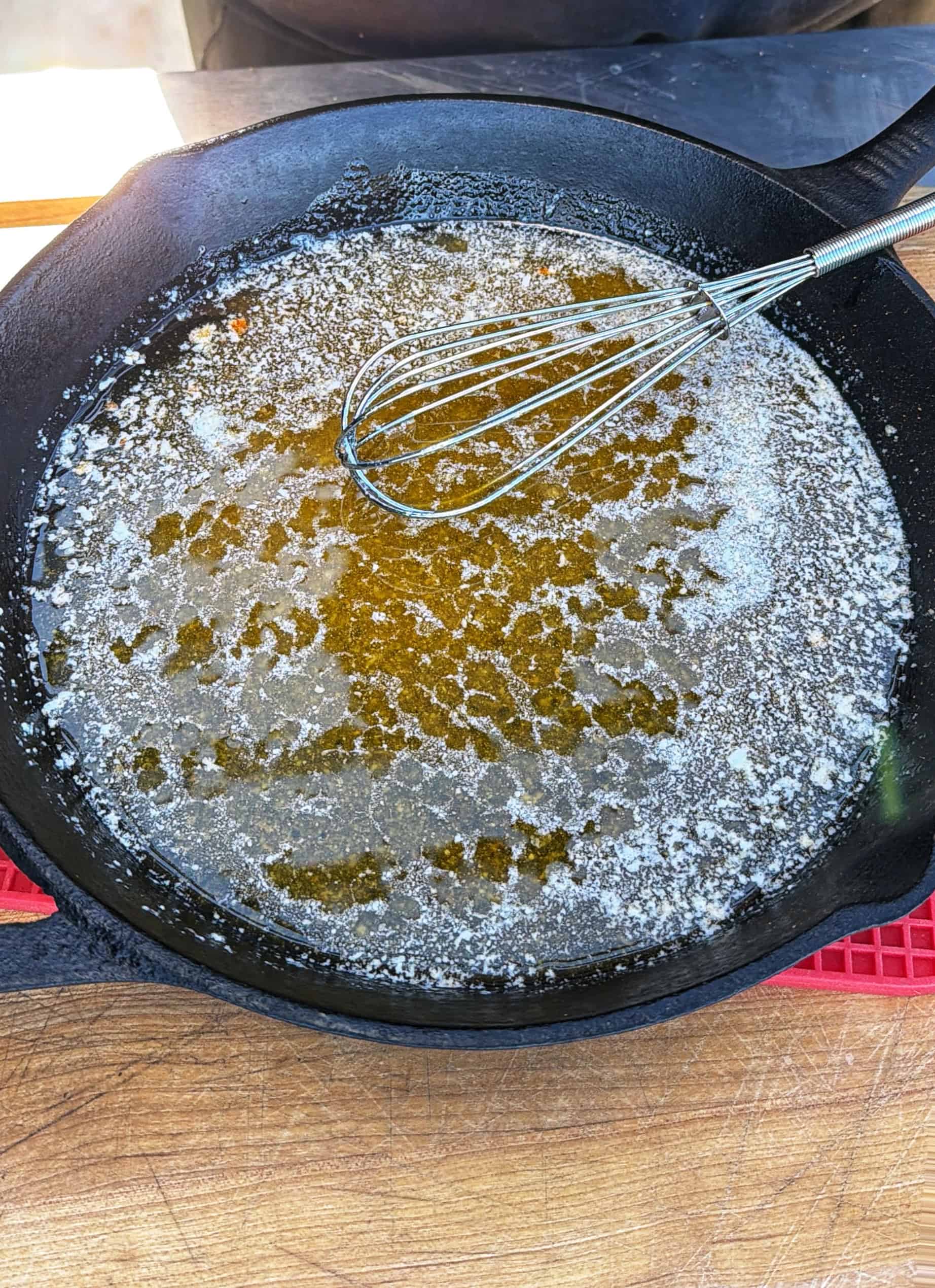 A metal whisk rests in a black cast iron skillet filled with melted butter, sitting on a wooden surface with a red silicone trivet partially visible underneath the pan.