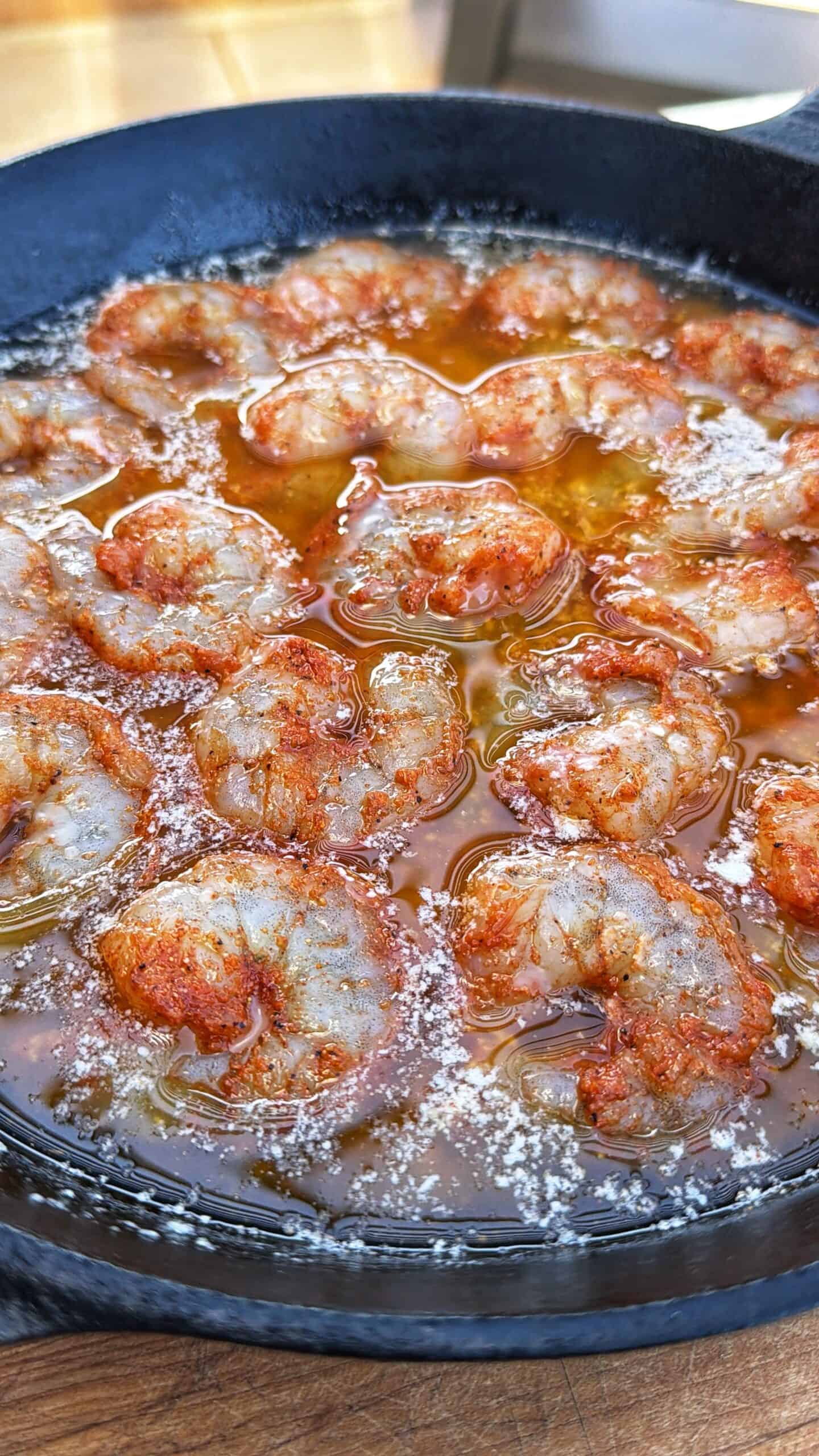 Shrimp cooking in sizzling oil in a black cast iron skillet, seasoned with spices and lightly dusted with salt, creating a golden, bubbling surface.