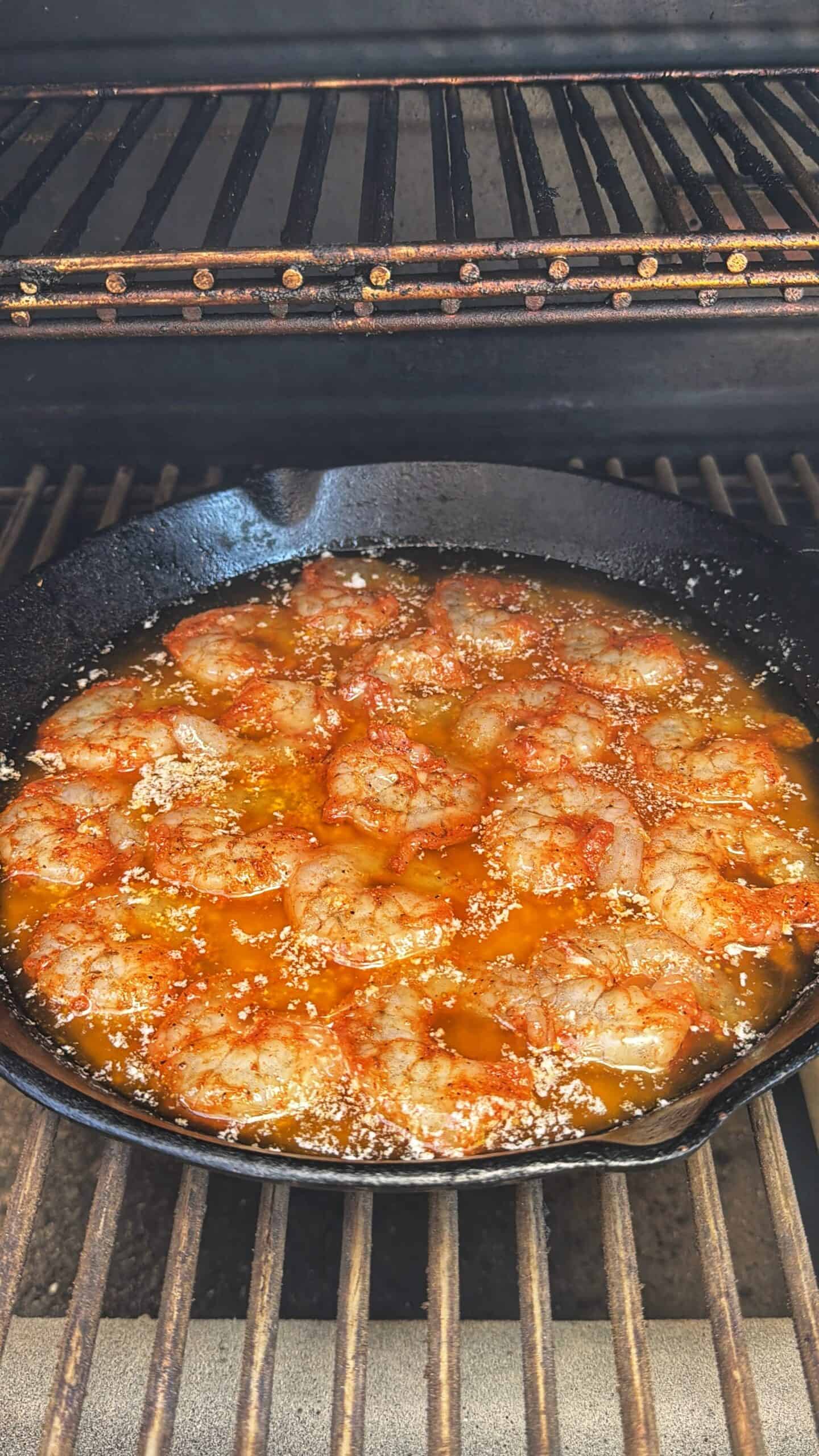 A cast iron skillet filled with shrimp in a seasoned, orange-colored sauce is cooking on a grill rack inside an open grill. The shrimp are topped with sesame seeds.