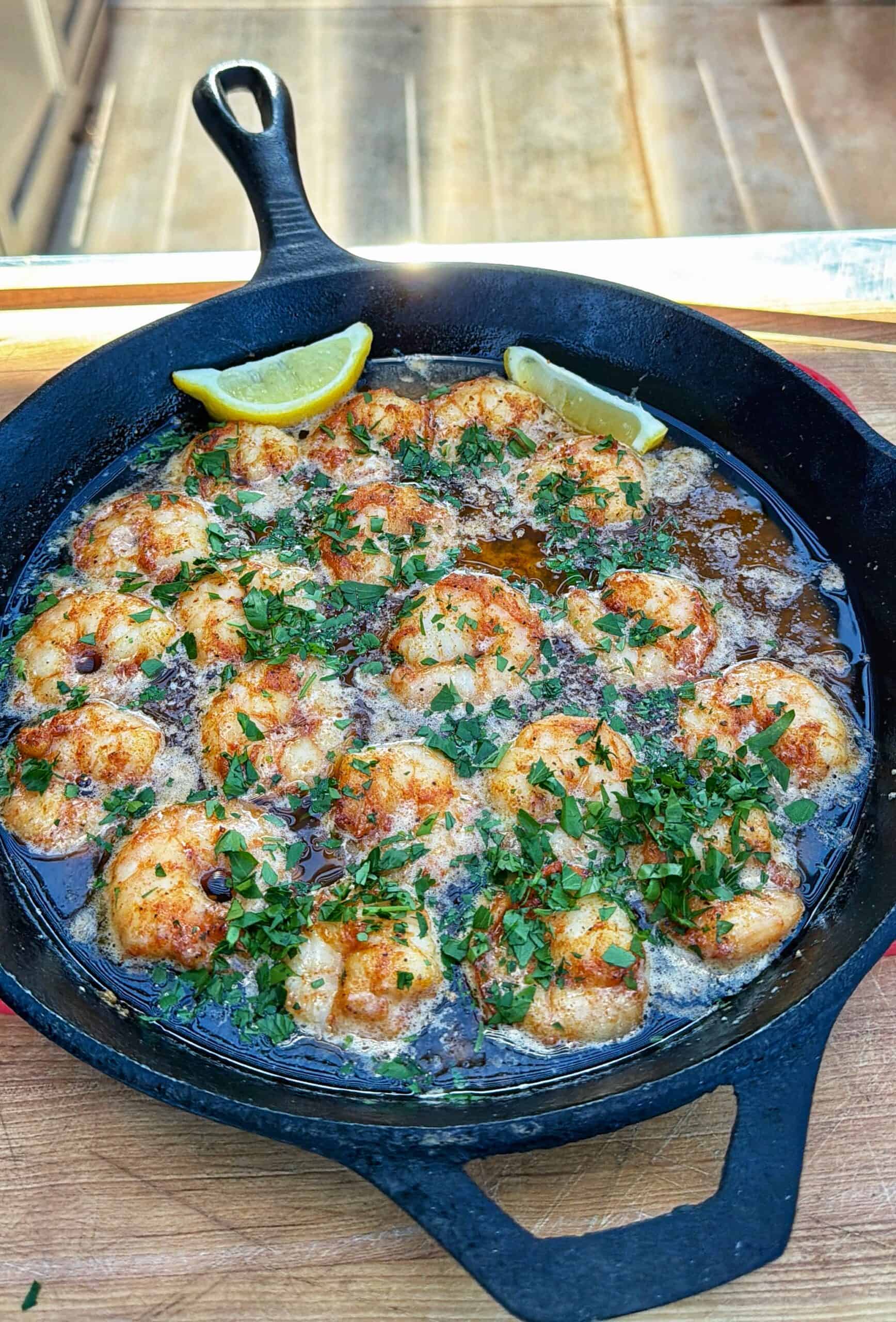 A cast iron skillet filled with shrimp cooked in a garlic butter sauce, garnished with chopped parsley and paprika, and served with two lemon wedges on the side.