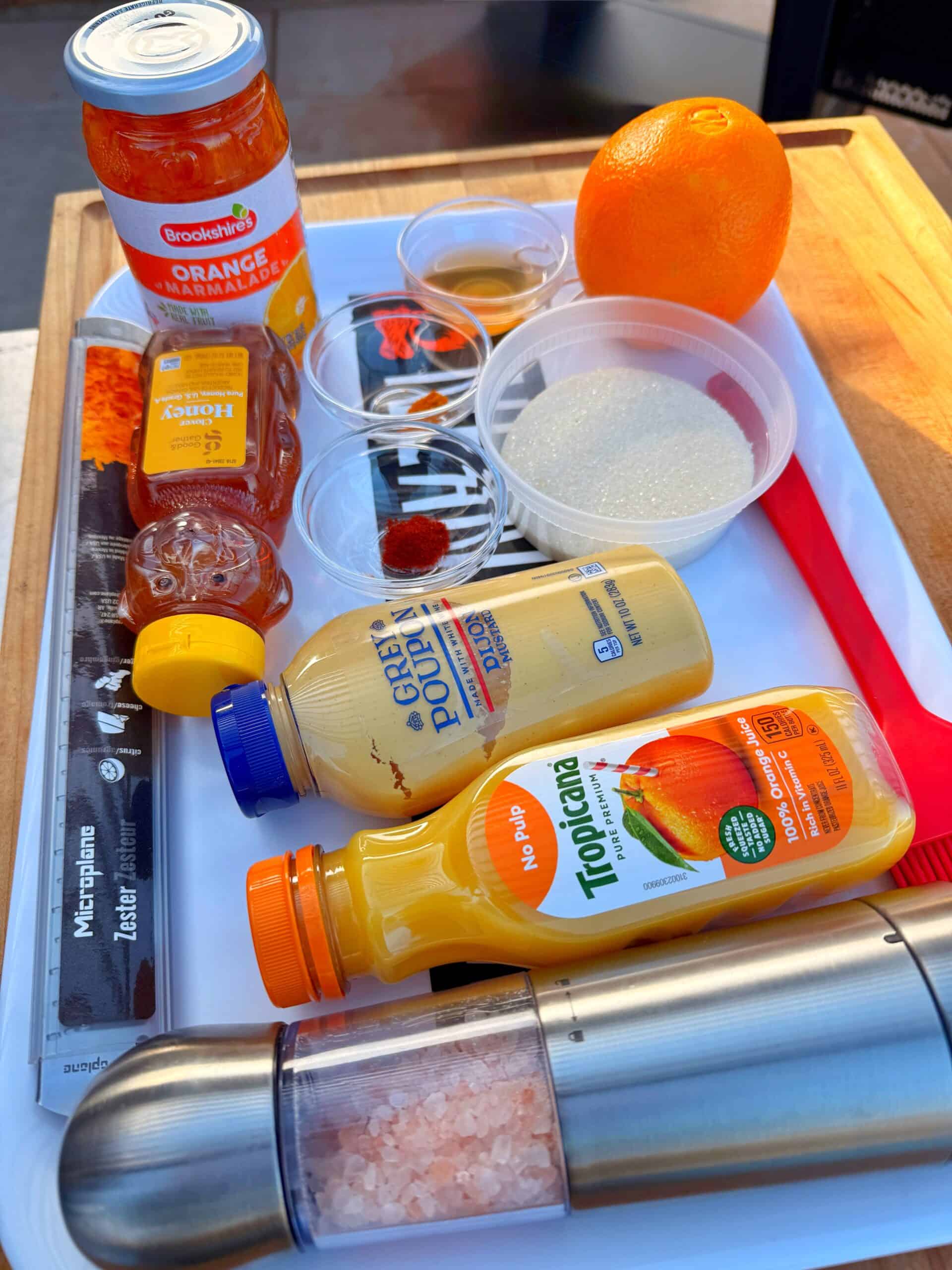 A tray with ingredients for a recipe, including a jar of orange marmalade, fresh orange, Tropicana orange juice bottles, Dijon mustard, honey, spices in small bowls, sugar, and a pink Himalayan salt grinder.