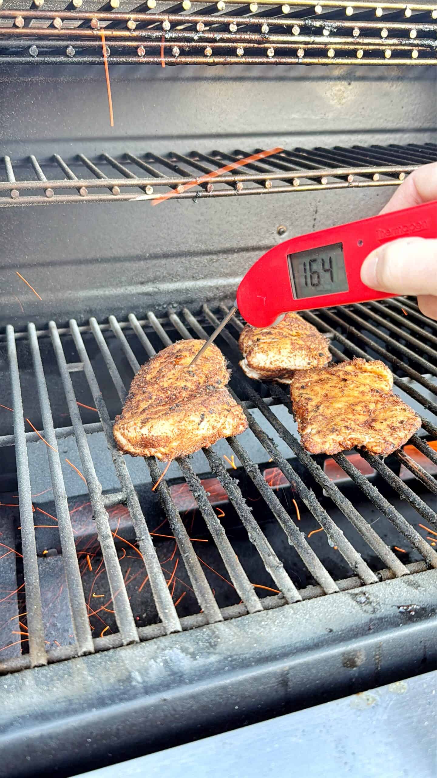 A hand holds a digital meat thermometer reading 164°F, checking the temperature of seasoned meat cooking on a grill. Three pieces of meat are on the grill grate with some orange sparks visible below.