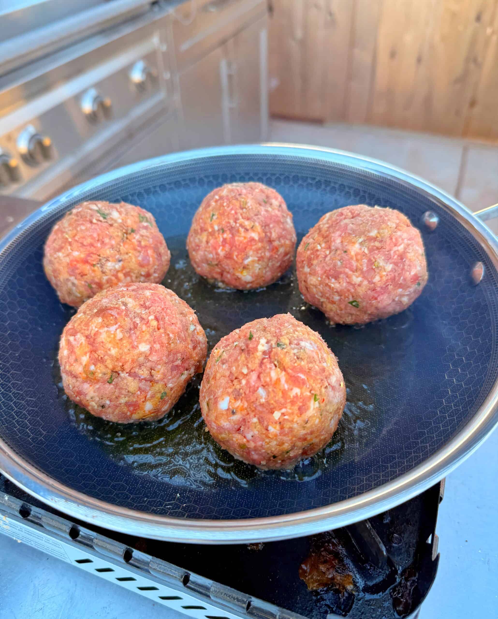 Five raw meatballs are cooking in a frying pan on an outdoor stove, with a stainless steel grill and wooden fence visible in the background.