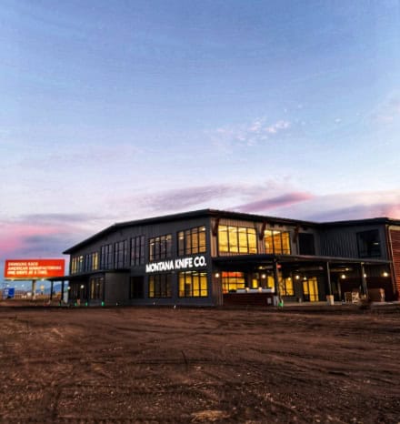 A large modern building with MONTANA KNIFE CO. lit up on the front stands under a colorful sky at sunset, surrounded by dirt and a mostly empty lot; a red signboard and hints of a Montana Knife Company review vibe complete the scene.