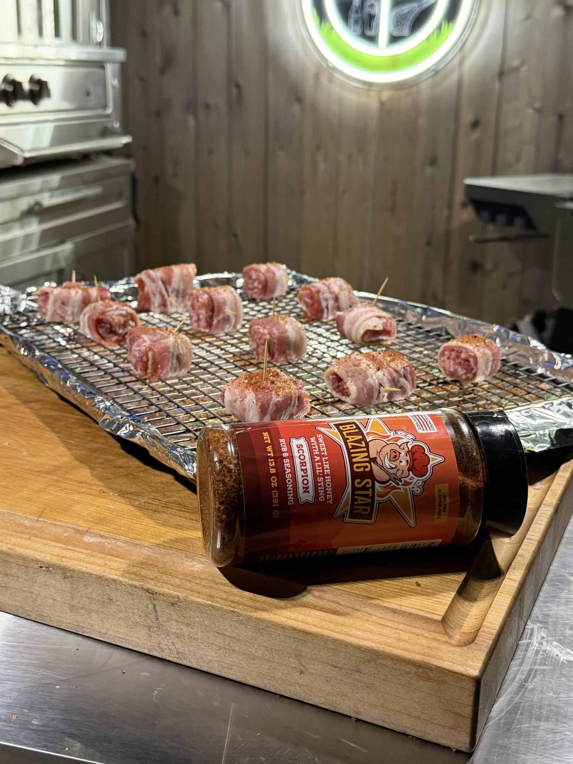 A tray of Bacon Wrapped Brat Bites rests on a foil-lined wire rack atop a wooden counter. In front, a bottle of Blazing Star barbecue seasoning lies on its side, with a grill visible in the background.