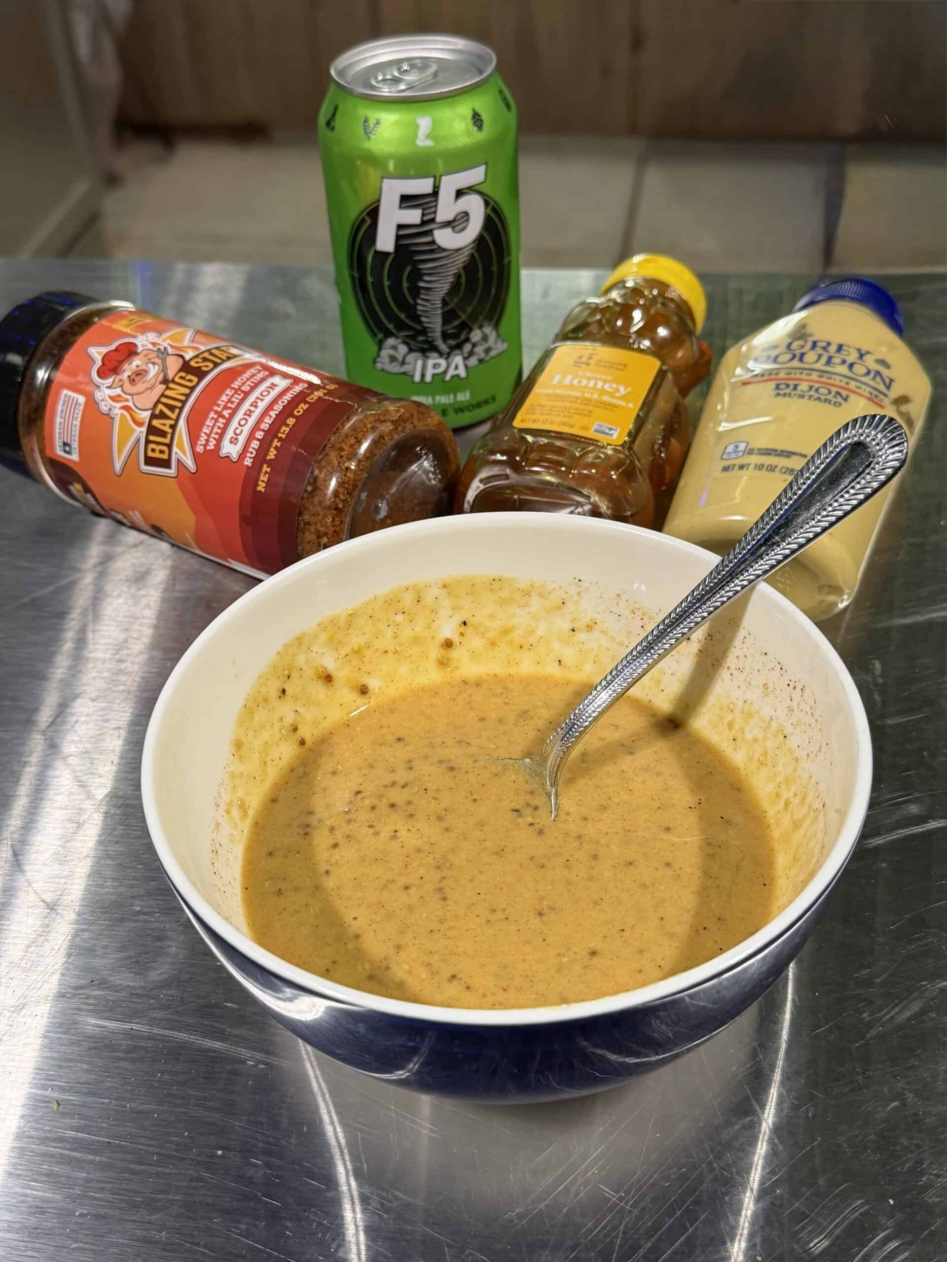 A bowl of seasoned sauce with a spoon inside sits on a metal surface. Behind it are a can of F5 IPA beer, a bottle of BBQ rub, honey, Dijon mustard, and ingredients ready for delicious Bacon Wrapped Brat Bites.
