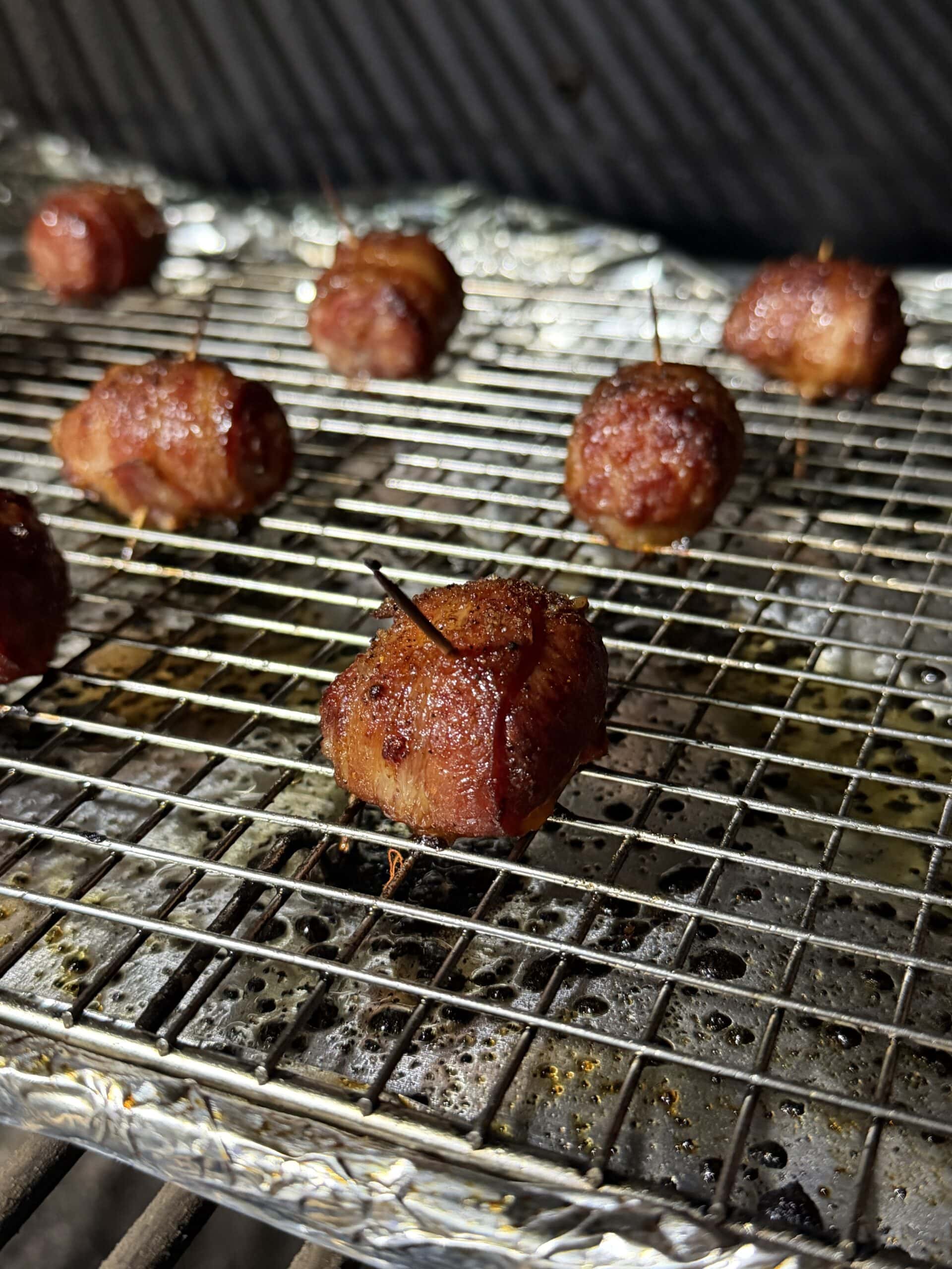 Bacon Wrapped Brat Bites appetizers secured with toothpicks are cooking on a metal rack lined with foil, with a few pieces in focus and several more blurred in the background.