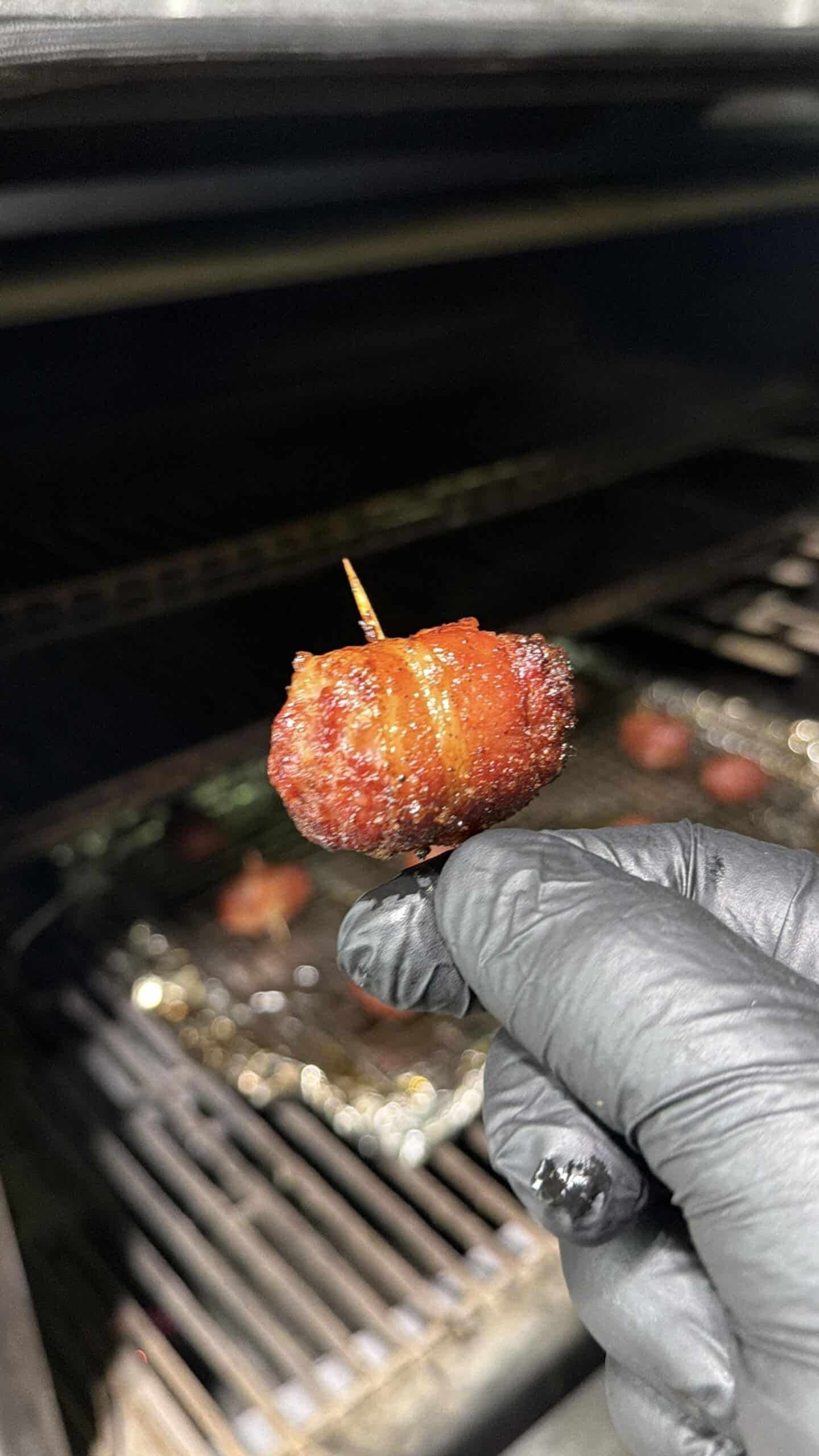 A gloved hand holds a Bacon Wrapped Brat Bite secured with a toothpick in front of a grill, with more Bacon Wrapped appetizers sizzling on a tray in the background.