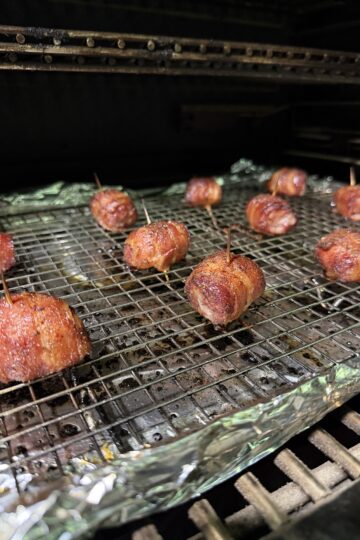 Bacon Wrapped Brat Bites, secured with toothpicks, are arranged on a wire rack over a foil-lined tray, cooking in the oven or smoker until the bacon appetizer is crisp and browned.