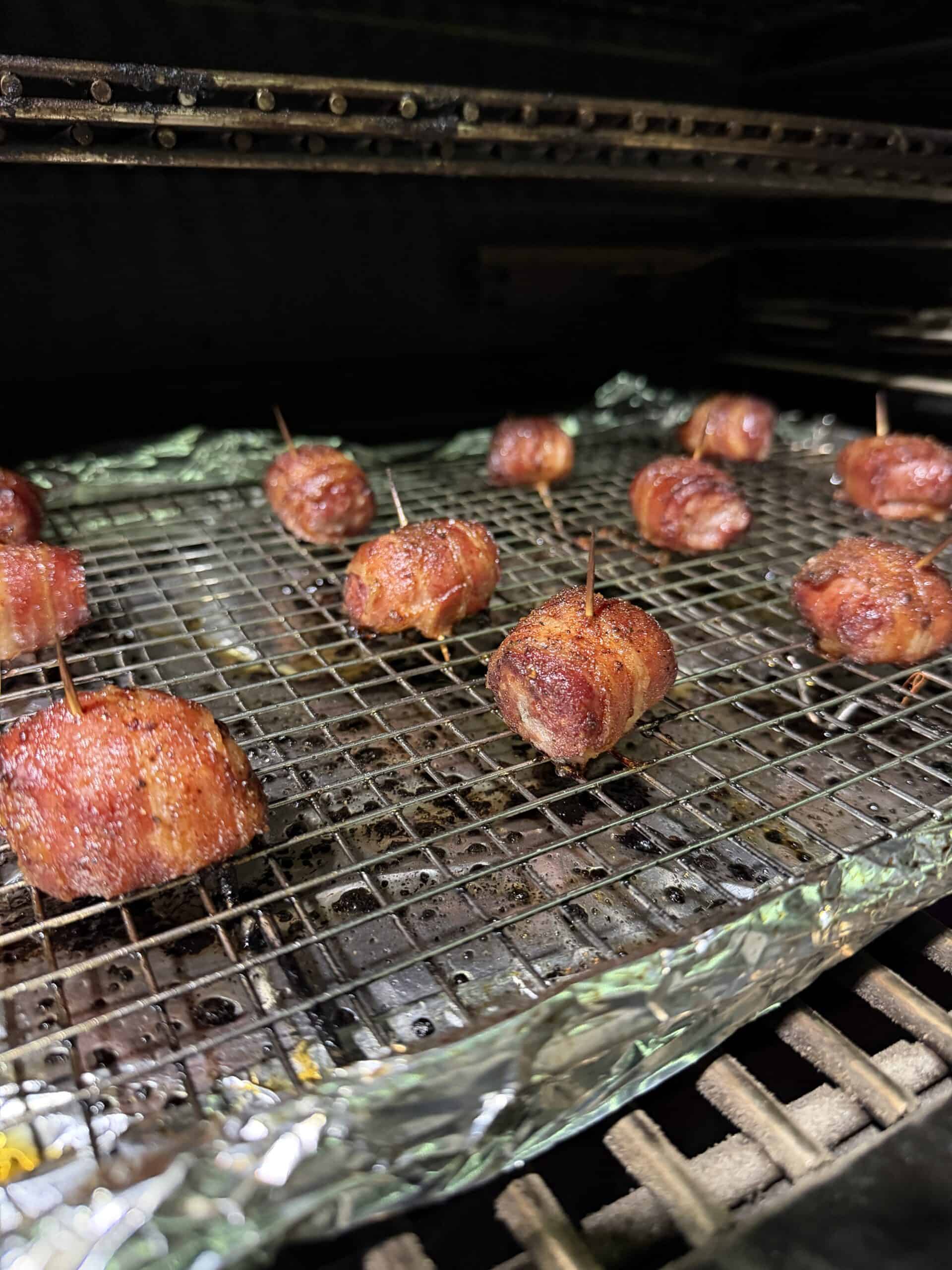 Bacon Wrapped Brat Bites, secured with toothpicks, are arranged on a wire rack over a foil-lined tray, cooking in the oven or smoker until the bacon appetizer is crisp and browned.