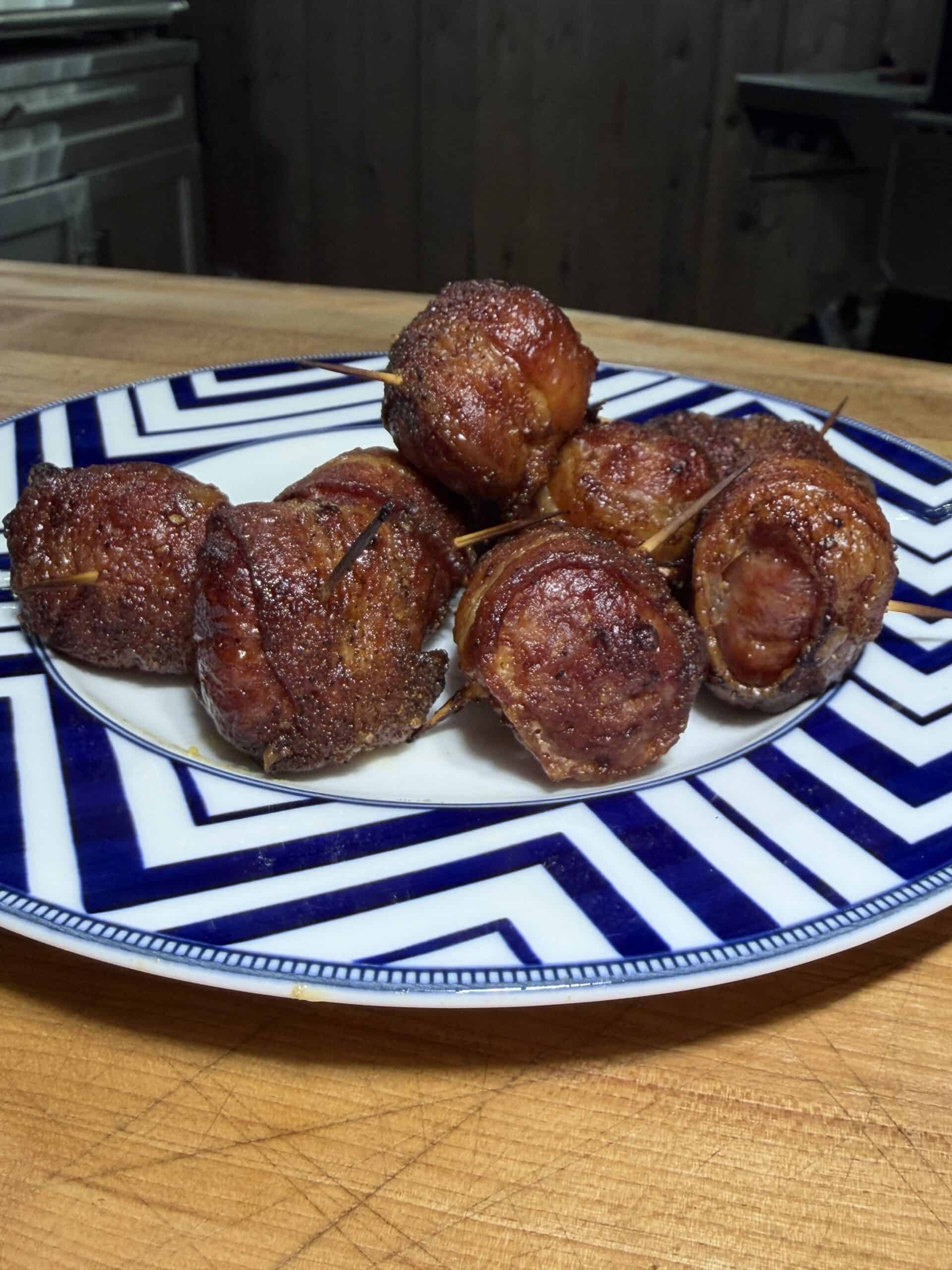 A plate of seven bacon-wrapped Brat Bites, secured with toothpicks, serves as a savory appetizer on a blue and white patterned dish atop a wooden countertop. Recipe is for bacon wrapped brats