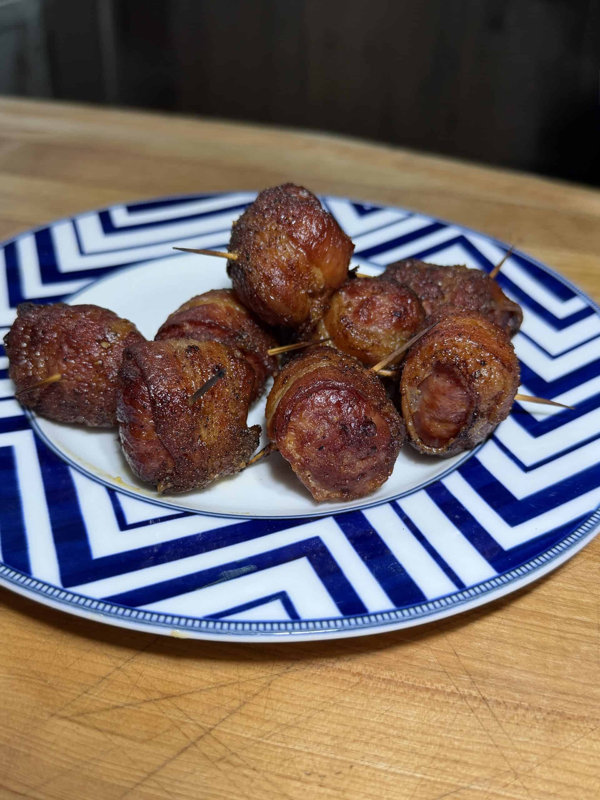 A plate of bacon wrapped brat bites, secured with toothpicks, sits on a blue and white patterned dish atop a wooden surface—an irresistible appetizer for any gathering.