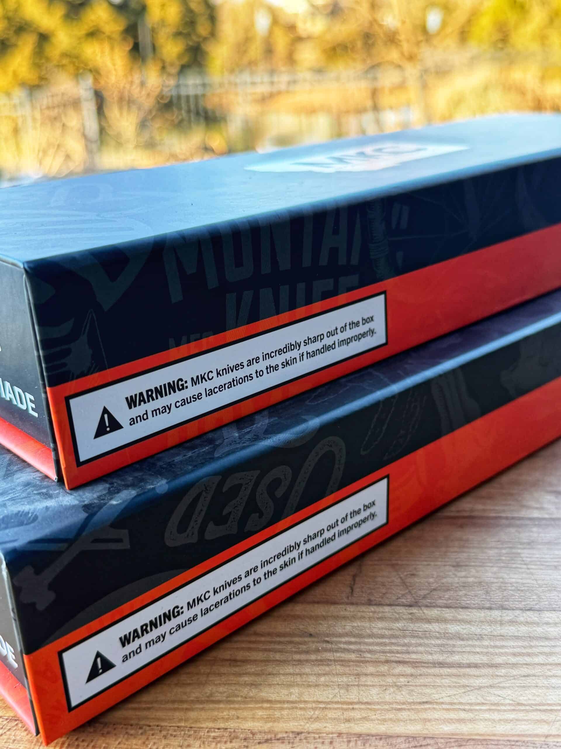 Two stacked knife boxes with a warning label that reads: MKC knives are incredibly sharp out of the box and may cause lacerations to the skin if handled improperly, on a wooden surface.