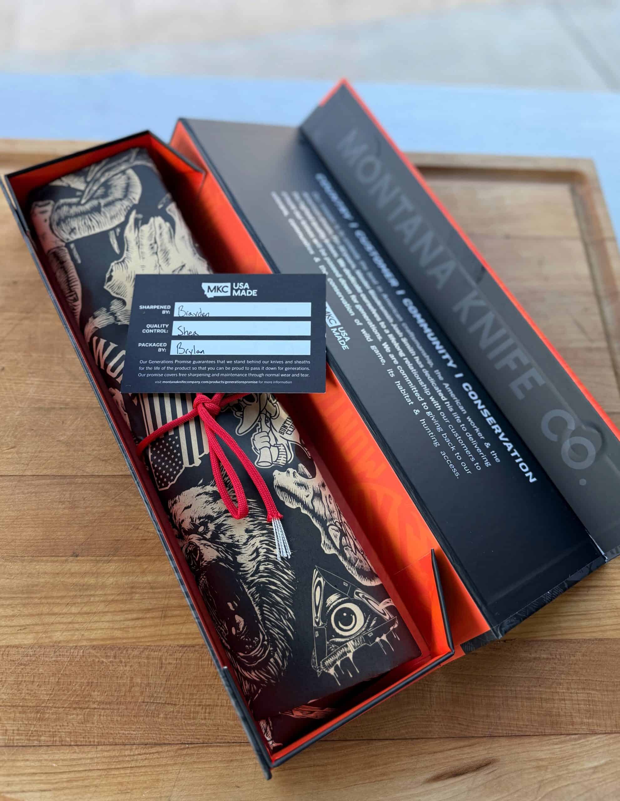 A Montana Knife Company box on a wooden surface holds a rolled knife wrap with animal designs, tied by a red string. An info card with handwritten details is placed on top.