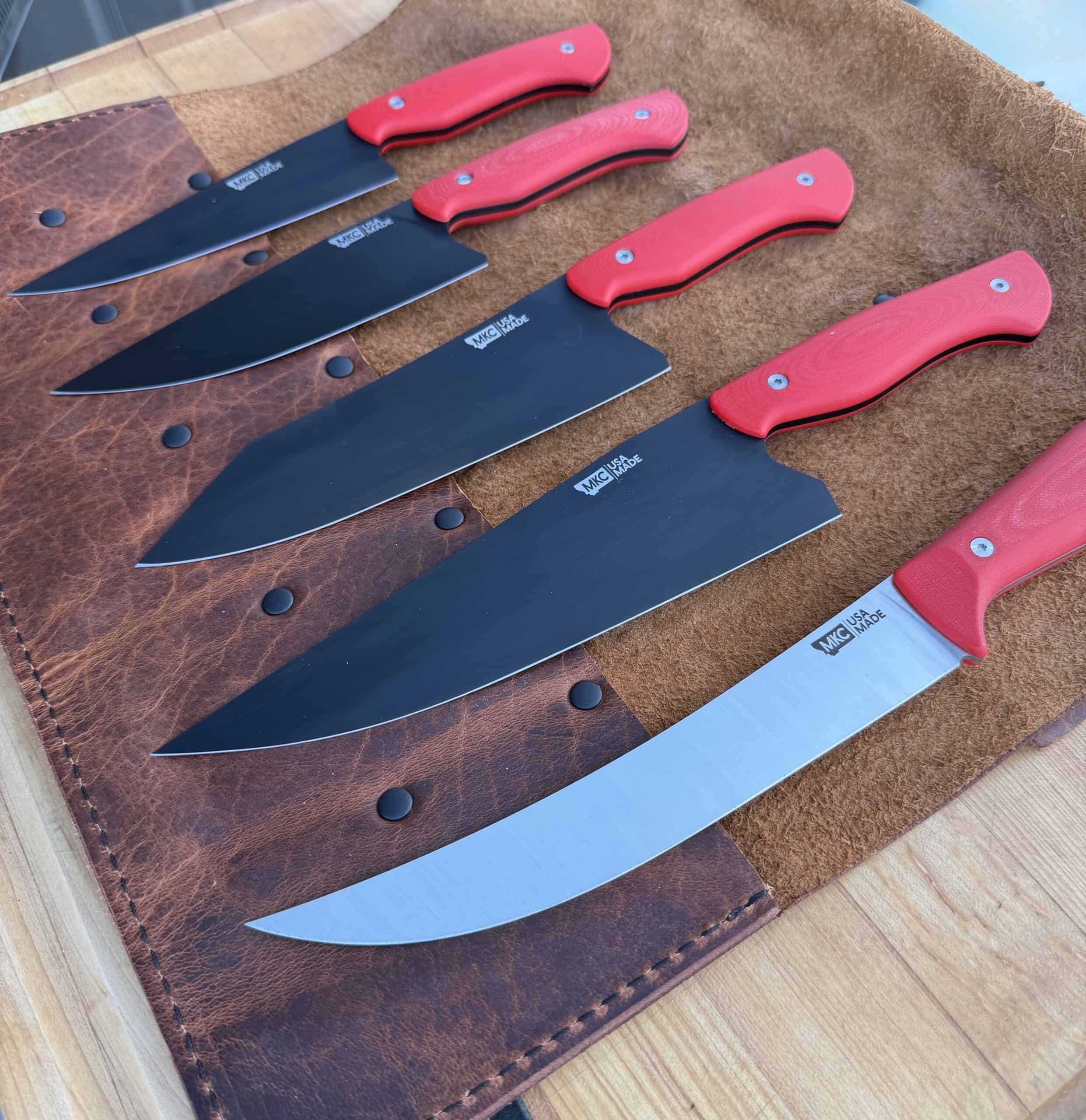 Five kitchen knives with red handles and black blades, except one with a silver blade, are neatly arranged on a brown leather knife roll on a wooden surface.