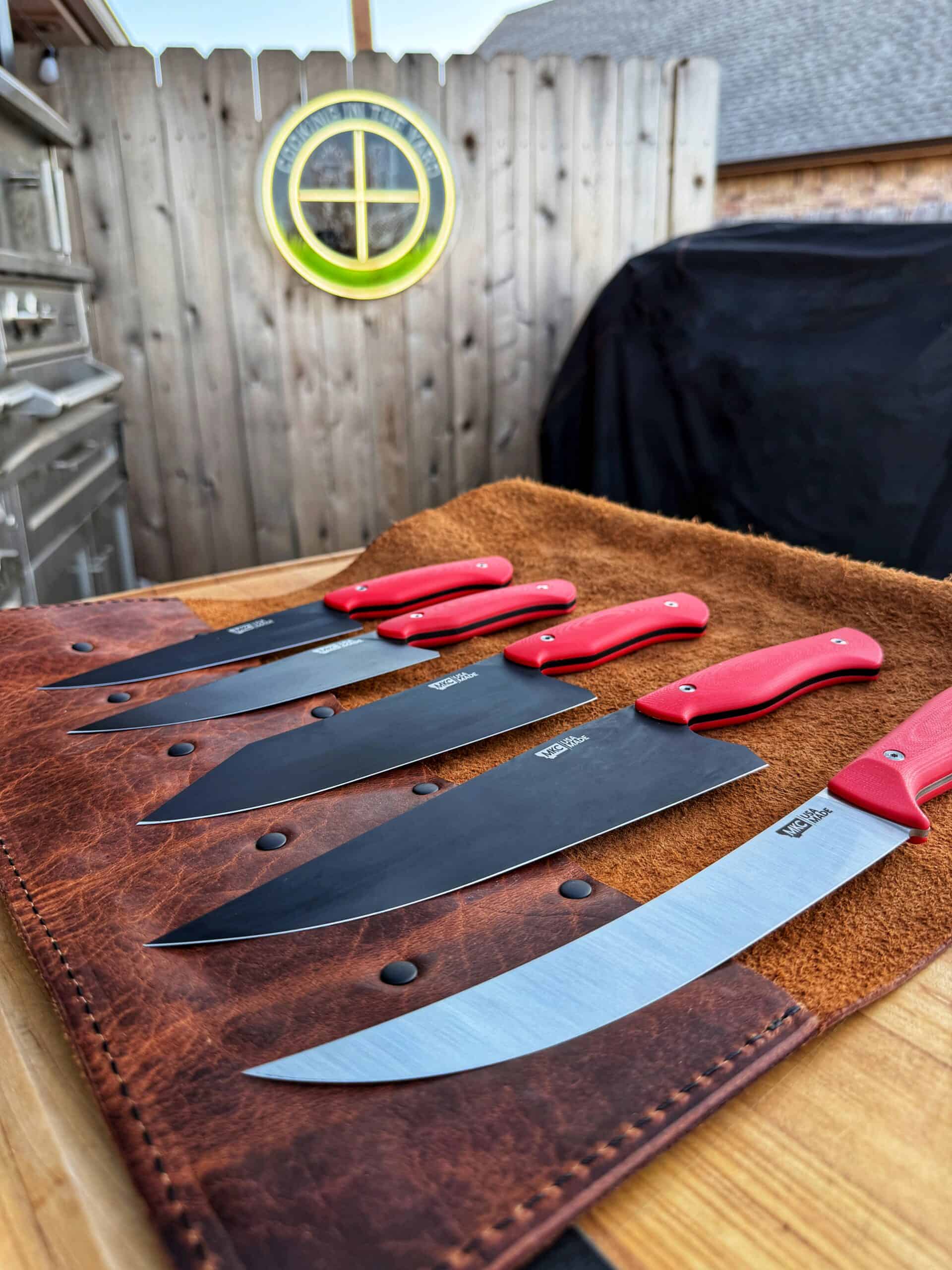 A set of five kitchen knives with red handles and black blades, arranged on a brown leather knife roll on an outdoor wooden table, with a wooden fence and house visible in the background.