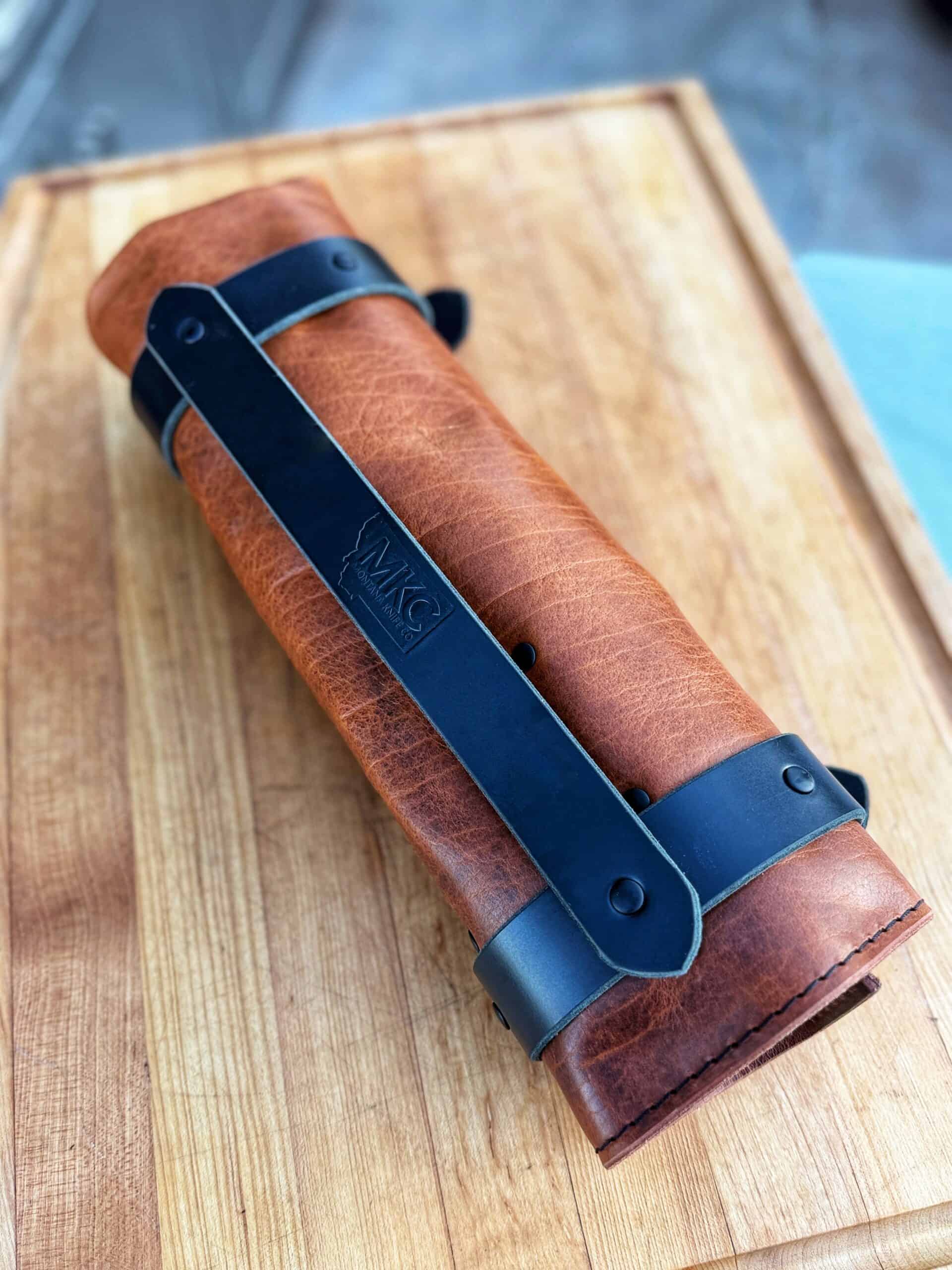 A rolled-up brown leather knife roll secured with black leather straps, resting on a wooden surface. The strap features an embossed logo that reads MKC.