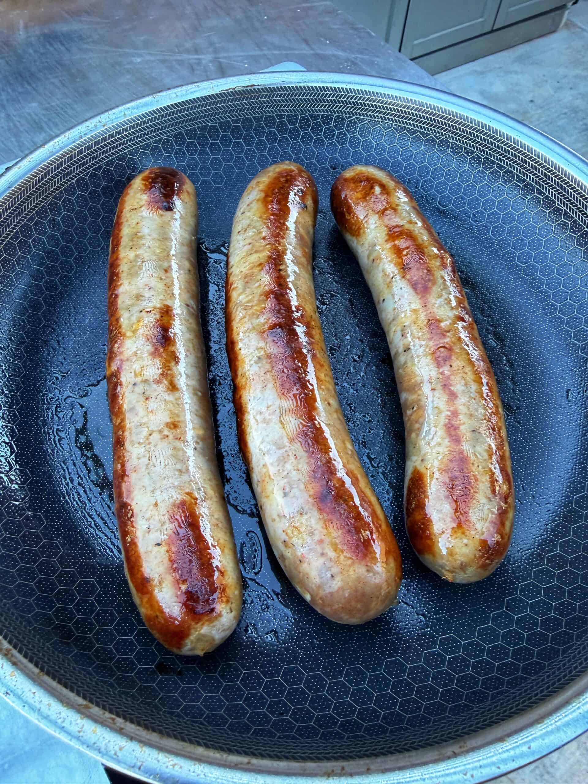 Three browned sausages are cooking in a nonstick skillet, with some oil visible around them. The sausages are evenly spaced and have a crispy, golden-brown surface.