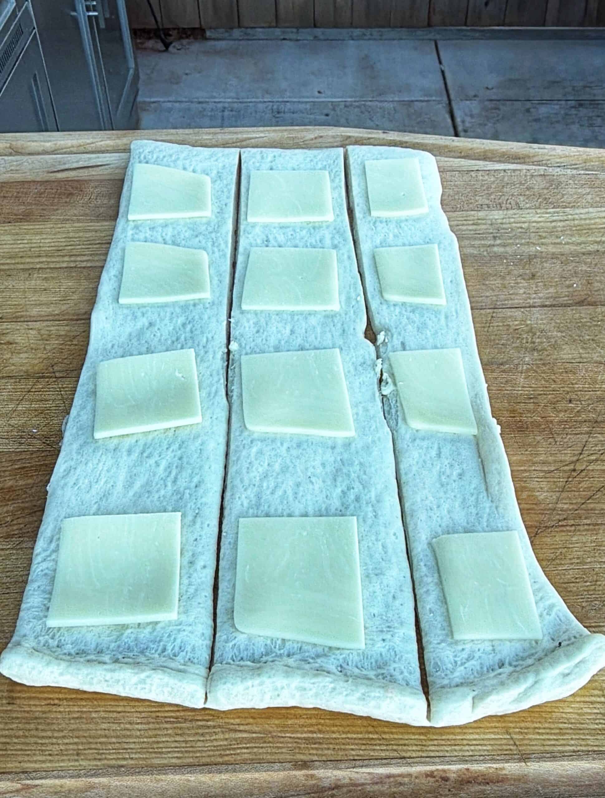 Rectangular dough is laid out on a wooden surface, cut into three long strips. Each strip is topped with evenly spaced square slices of white cheese. A concrete floor and cabinets are visible in the background.