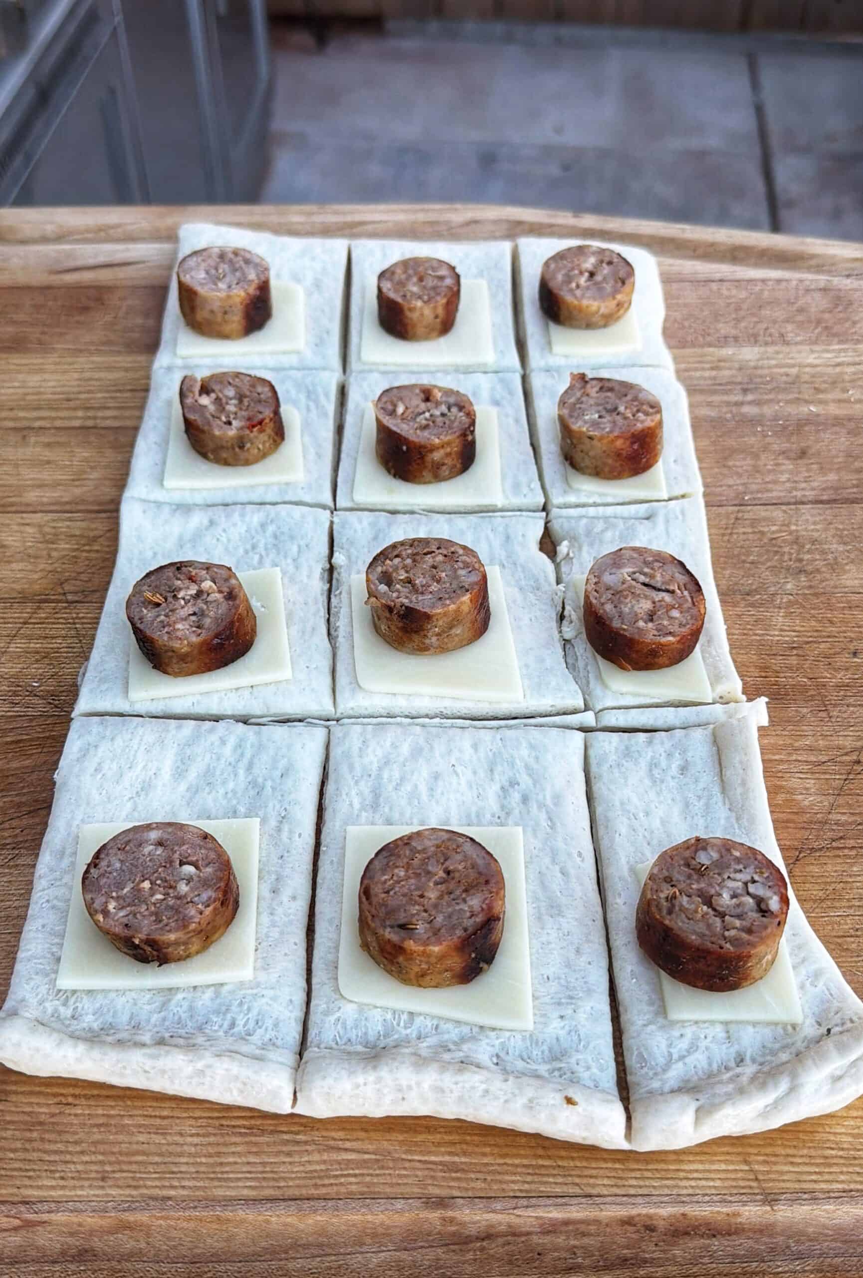 Rectangles of dough are laid out on a wooden surface, each topped with a square slice of cheese and a piece of sausage, arranged in a grid pattern.