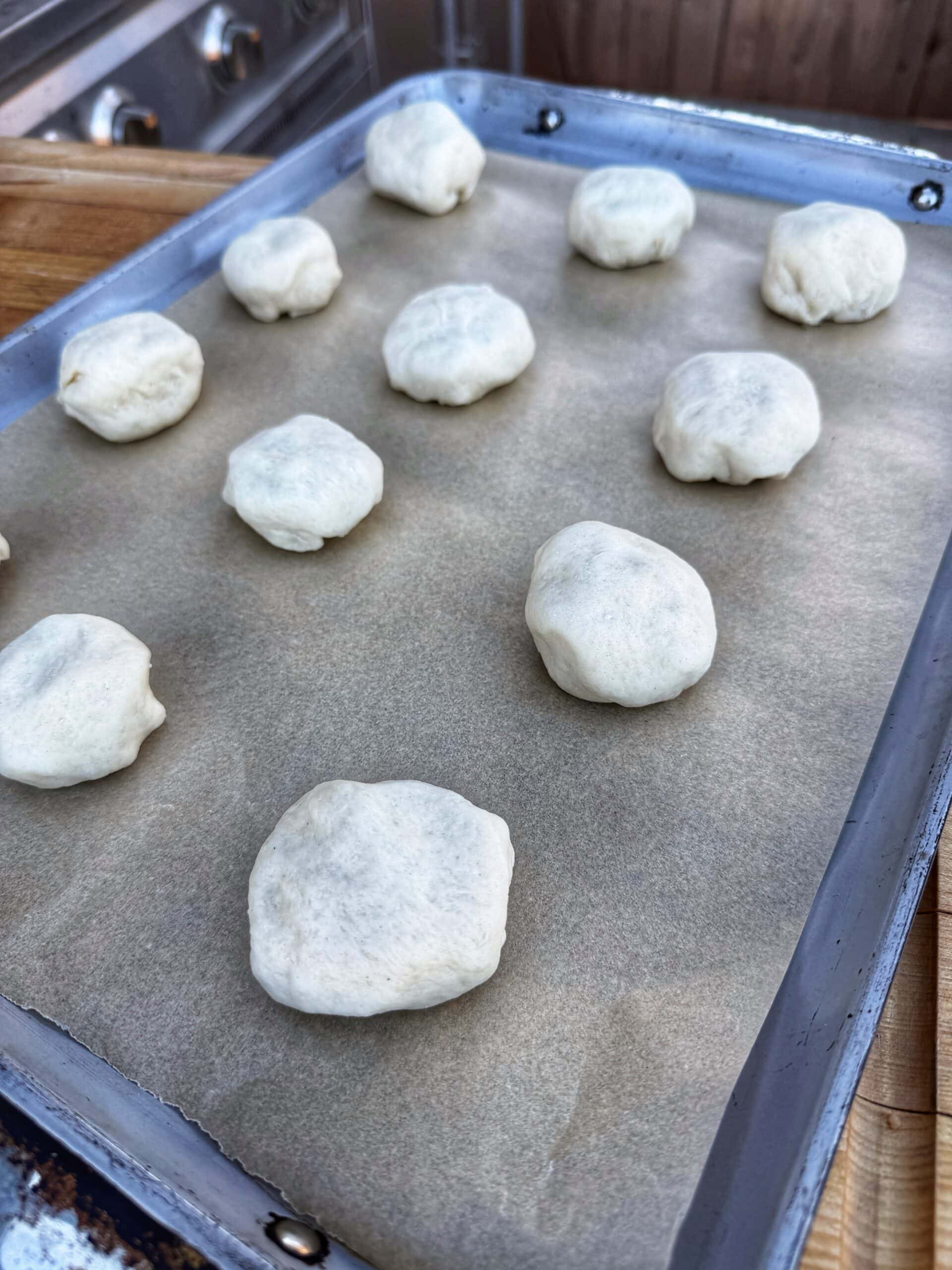 A baking tray lined with parchment paper holds several evenly spaced balls of raw dough, ready to be baked. The tray is set on a wooden surface in a kitchen.
