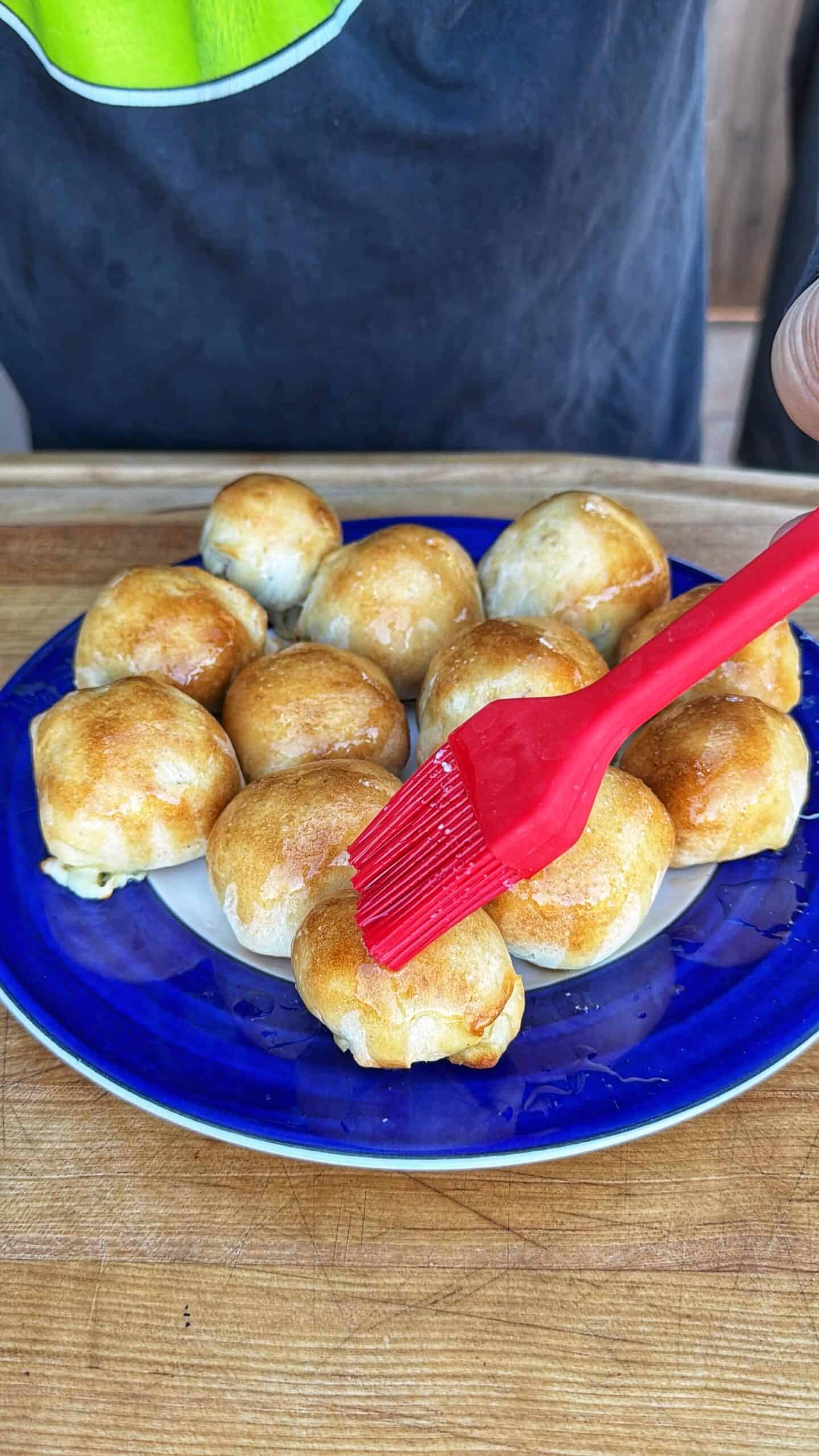 A person uses a red brush to glaze small, golden-brown baked rolls arranged on a blue plate placed on a wooden surface.