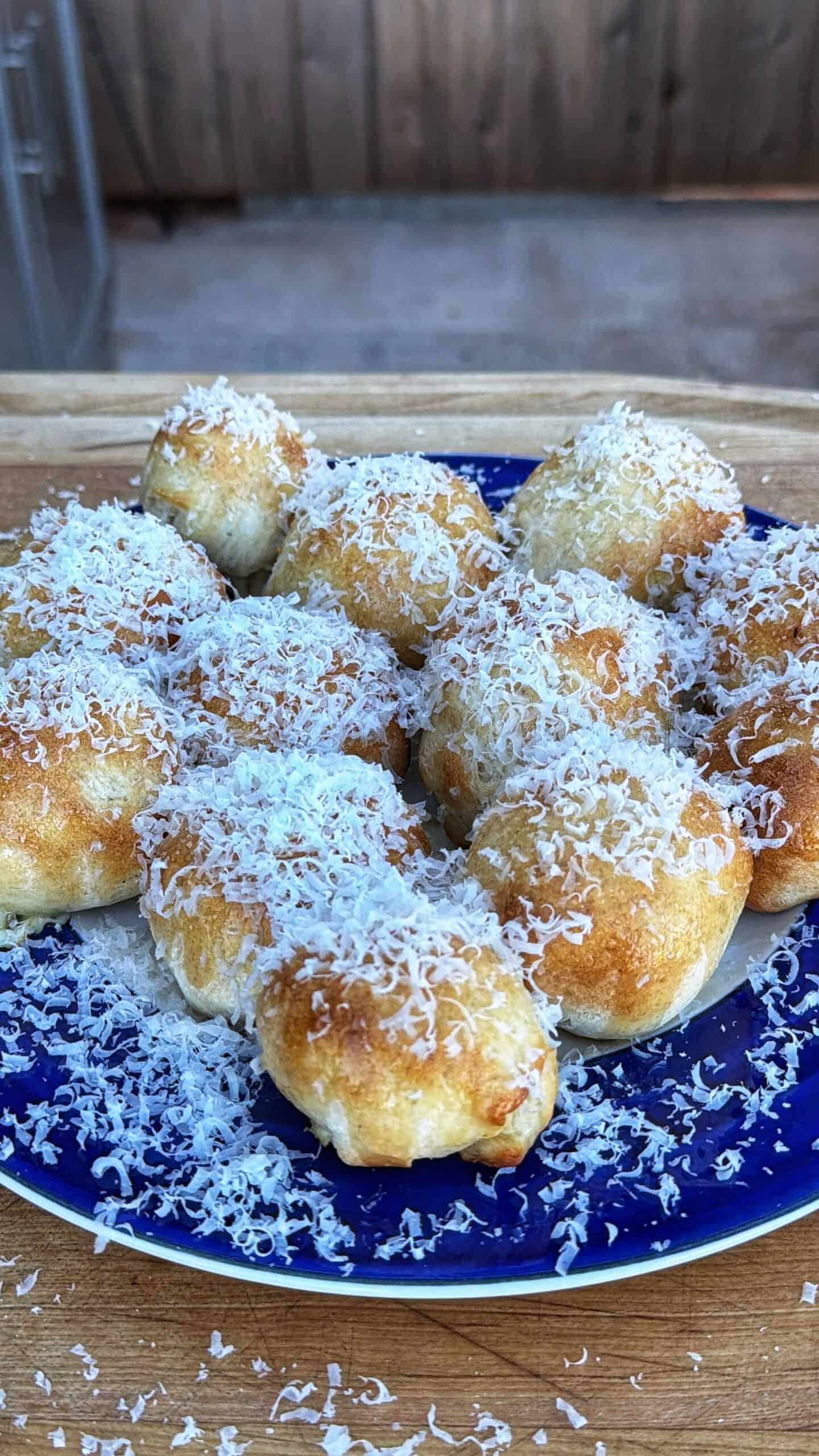 A blue plate holds a dozen golden-brown, round pastries topped generously with finely grated cheese, sitting on a wooden surface with a blurred background.