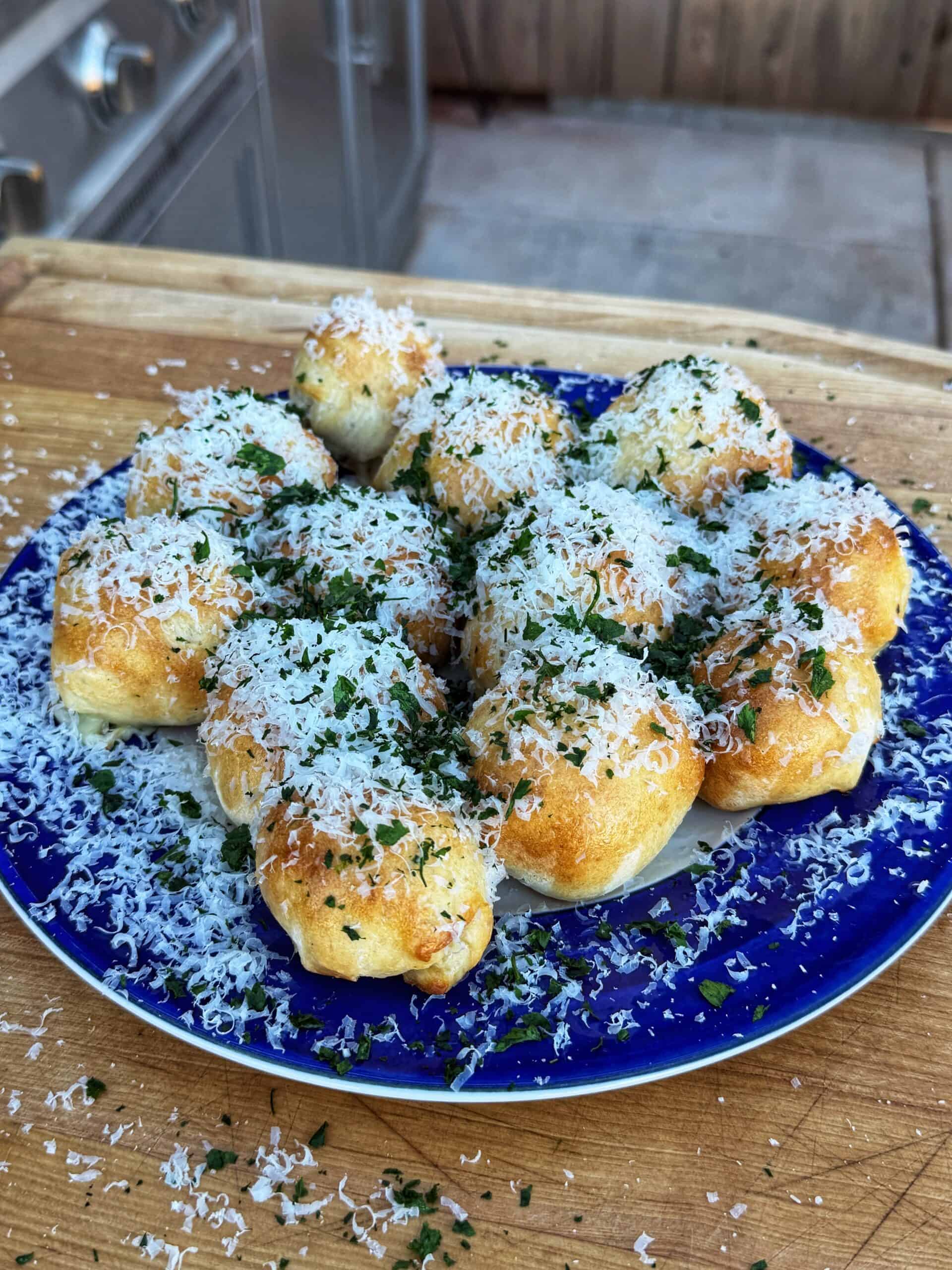 A blue plate holds golden, baked stuffed pizza bombs topped with shredded cheese and chopped herbs, sitting on a wooden surface outdoors.