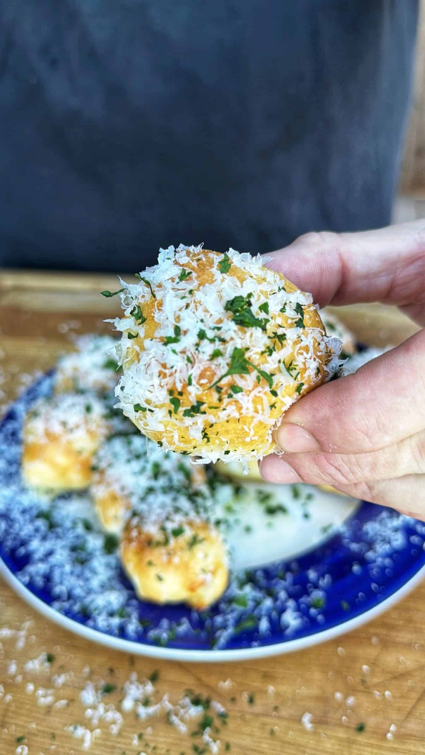 A hand holds up a small, round bread bite topped with grated cheese and chopped herbs. More cheesy bread bites are on a blue and white plate in the background.