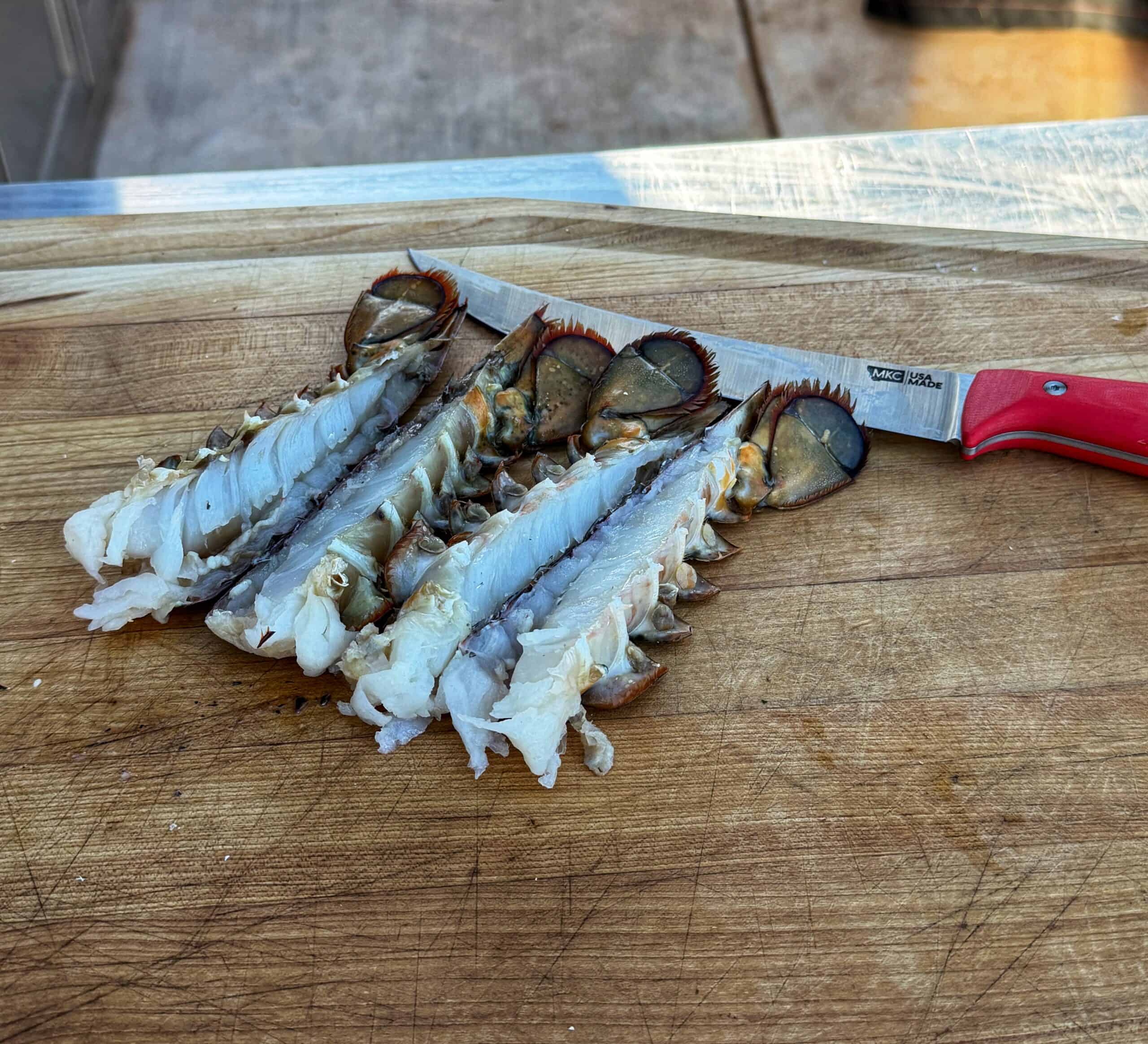 Four raw lobster tails split in half lie on a wooden cutting board with a knife that has a red handle. The lobster tails are neatly arranged in a row, displaying their white flesh.