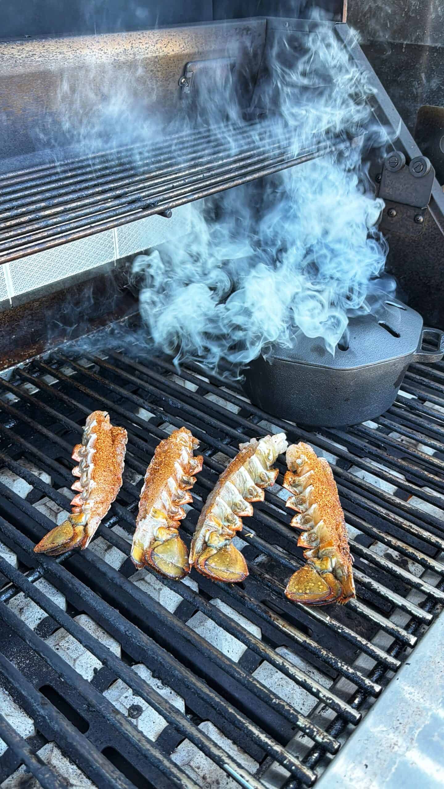 Four lobster tails are grilling on a barbecue next to a black cast iron pot, with smoke rising and partially filling the air above the grill.