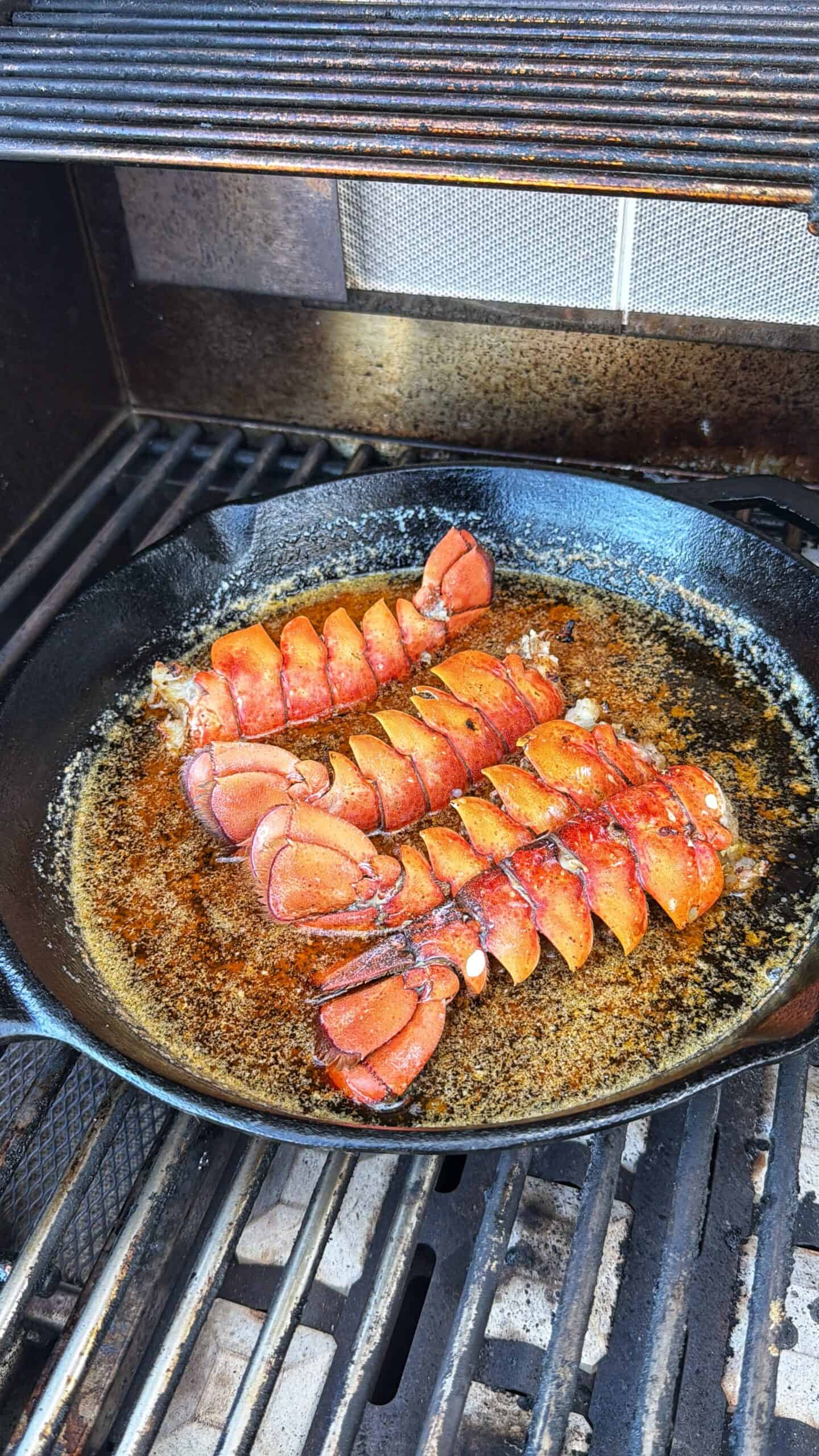 Four lobster tails are cooking in a cast iron skillet with melted butter on a grill, with the lobster shells facing up and the grill bars visible below.