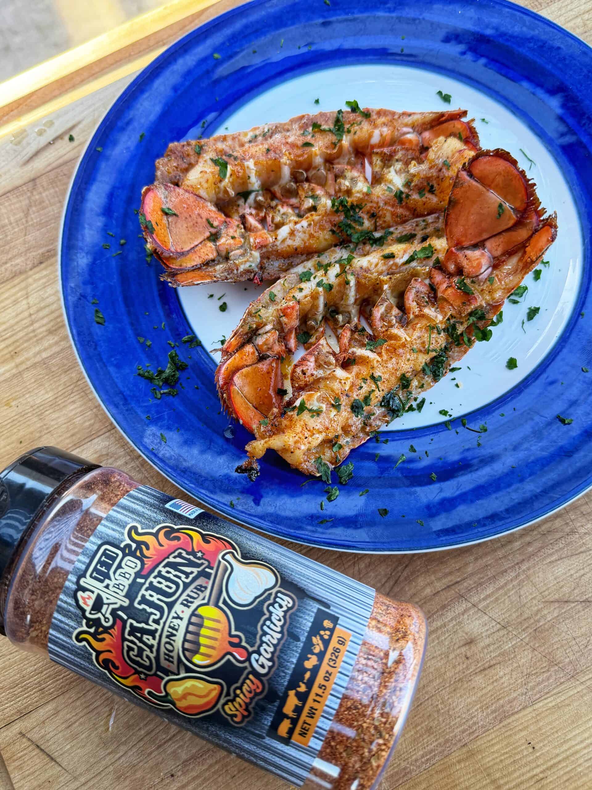Two smoked lobster tails garnished with chopped herbs on a blue-rimmed white plate, with a bottle of Cajun seasoning labeled Smokin Ghost Garlic placed nearby on a wooden surface.  This is a smoked lobster recipe