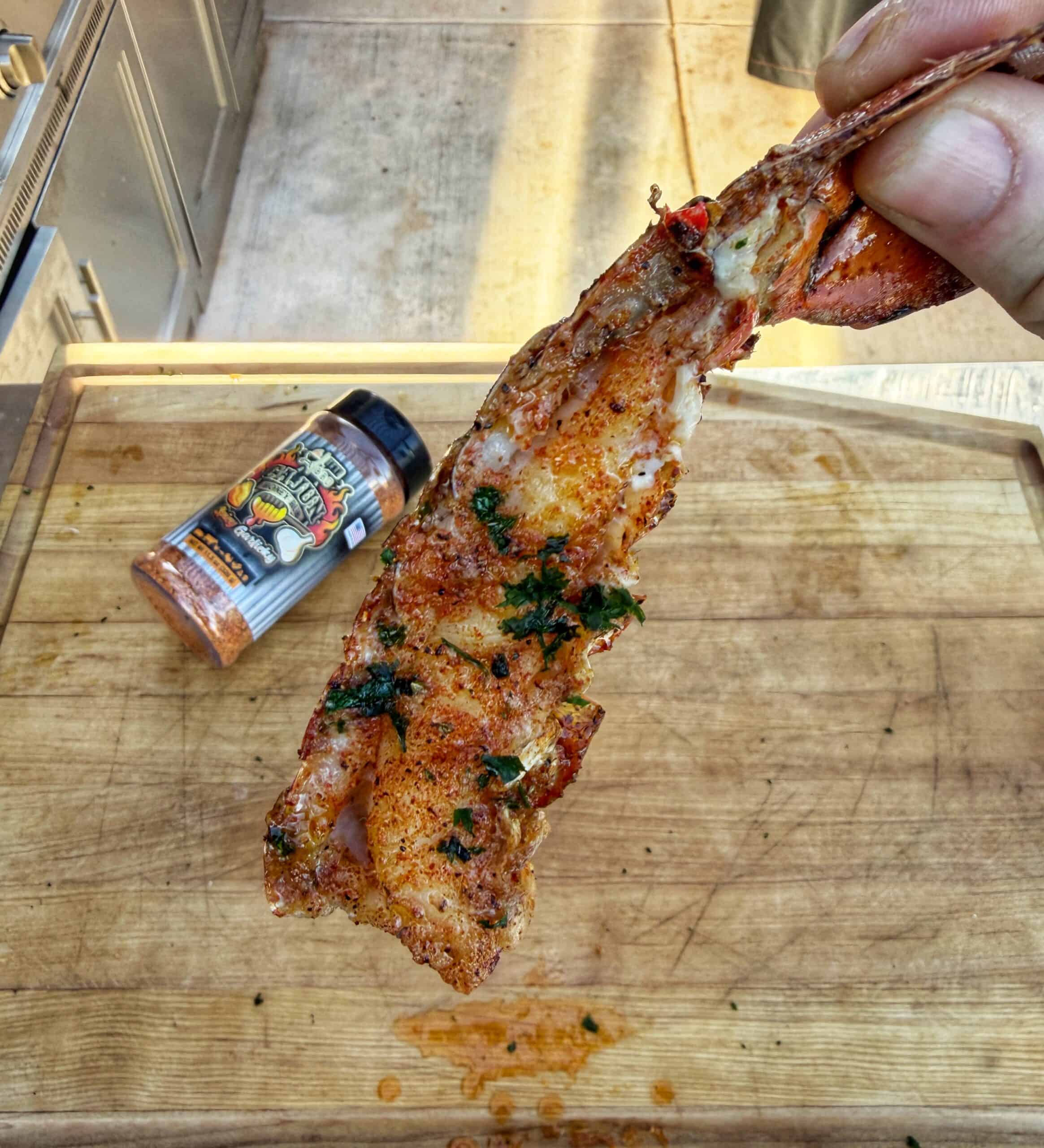 A hand holds up a grilled, seasoned lobster tail over a wooden cutting board. A container of spice mix lies on the board in the background, slightly out of focus.