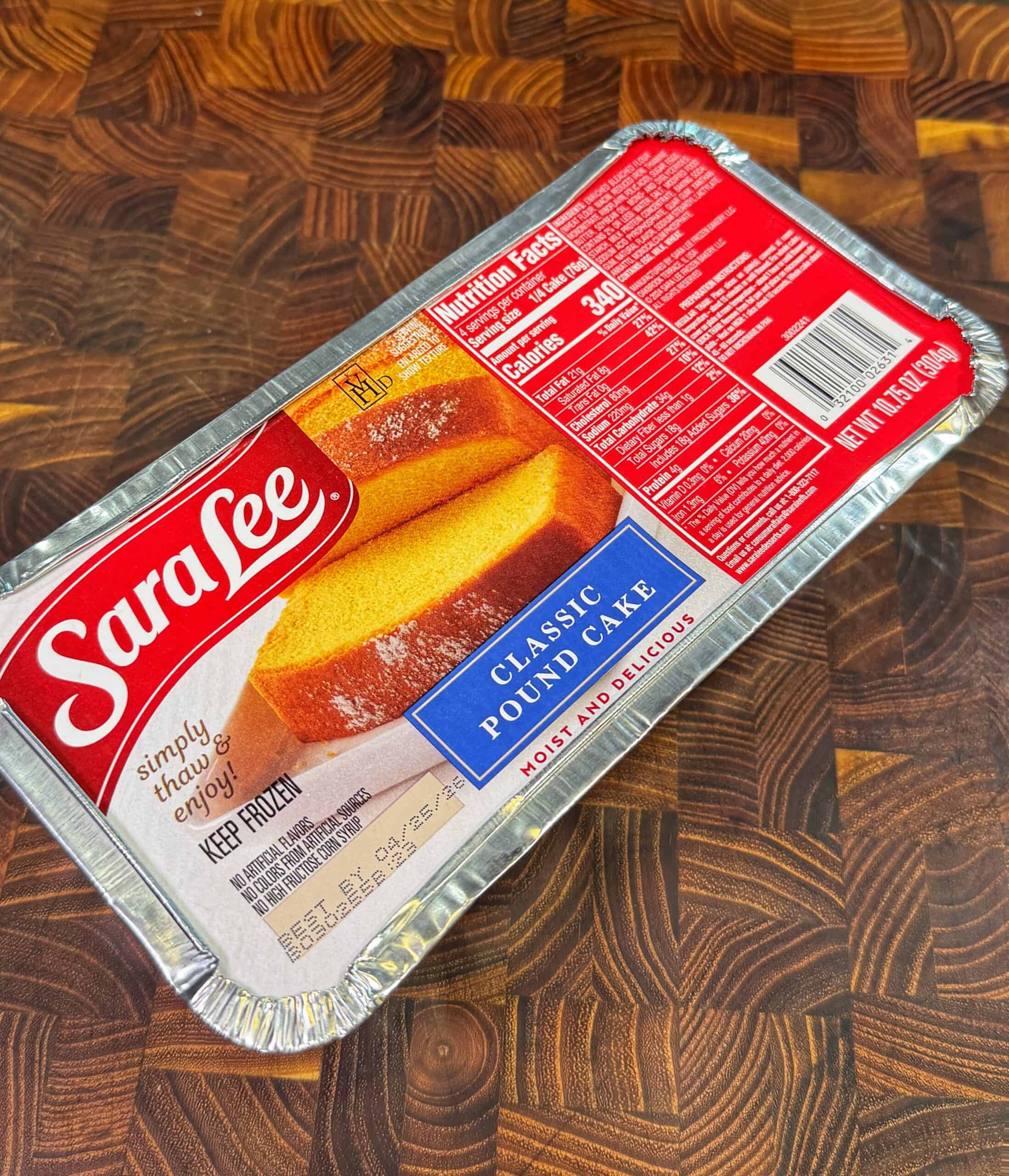 A Sara Lee Classic Pound Cake in a foil tray sits on a wooden countertop. The packaging shows an image of the cake, nutrition facts, and the brand logo in red and white.