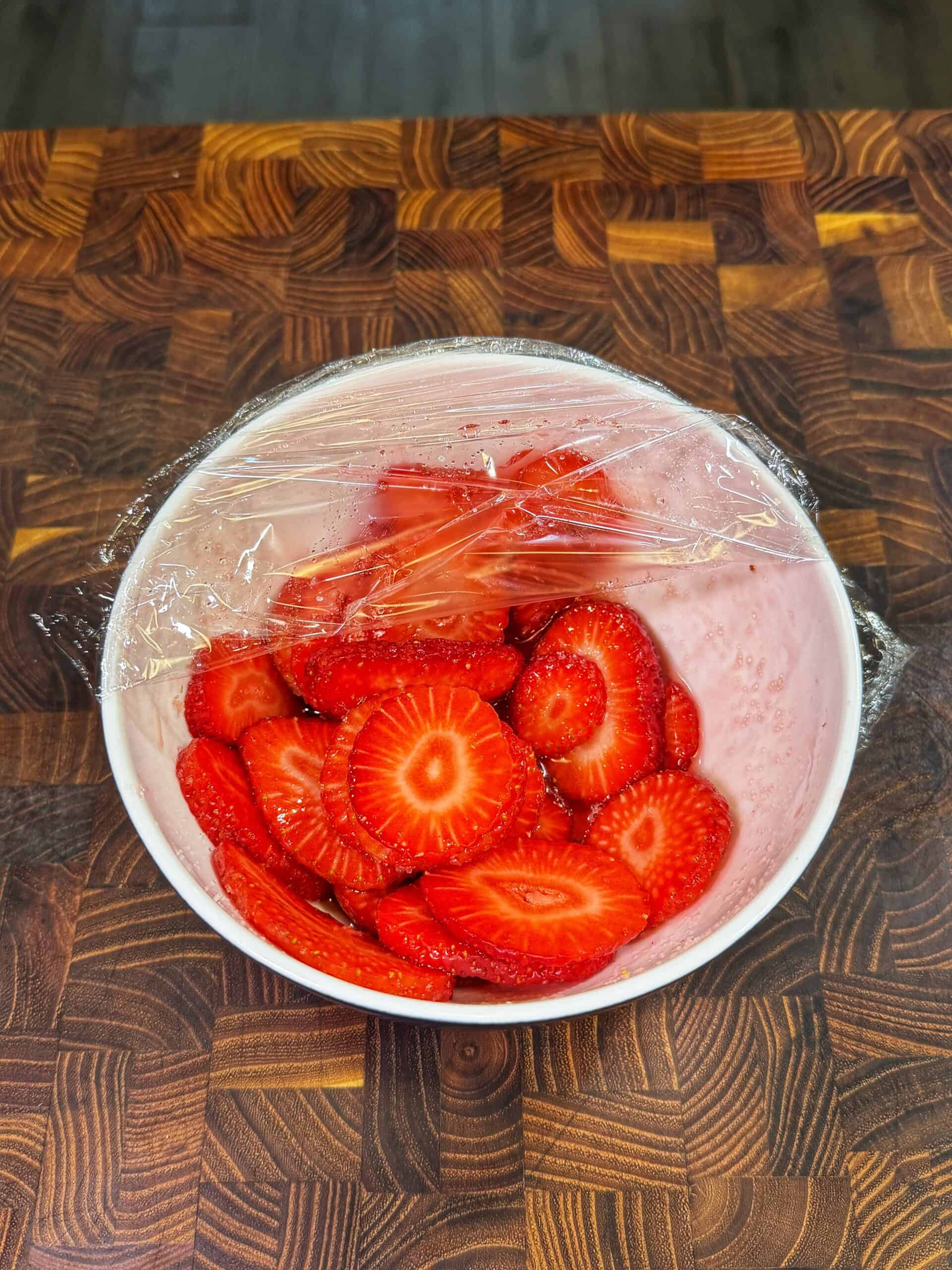 A white bowl filled with sliced strawberries is covered with a sheet of plastic wrap. The bowl sits on a wooden countertop with a patterned design.