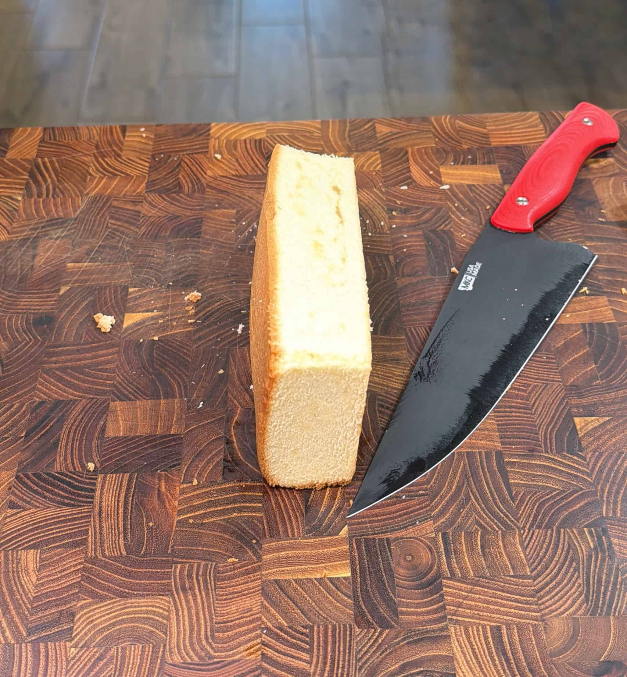 A loaf of bread standing upright on a wooden cutting board next to a large kitchen knife with a red handle. The board shows a distinctive geometric wood pattern.