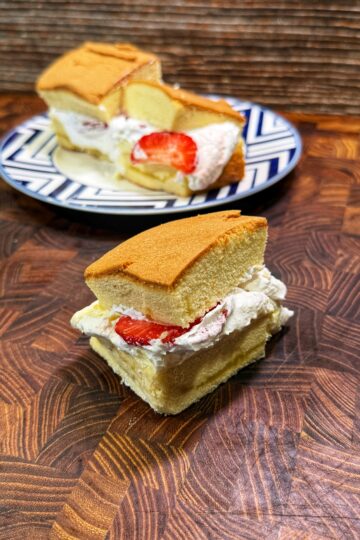 A slice of sponge cake filled with whipped cream and a strawberry sits on a wooden surface, with a plate holding another slice in the background.