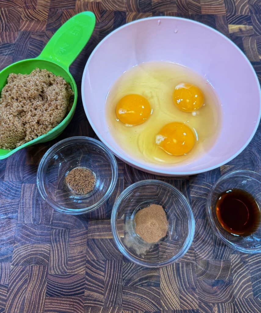 A pink bowl with three cracked eggs, a green measuring cup filled with brown sugar, and four small glass bowls containing spices and vanilla extract on a wooden countertop.